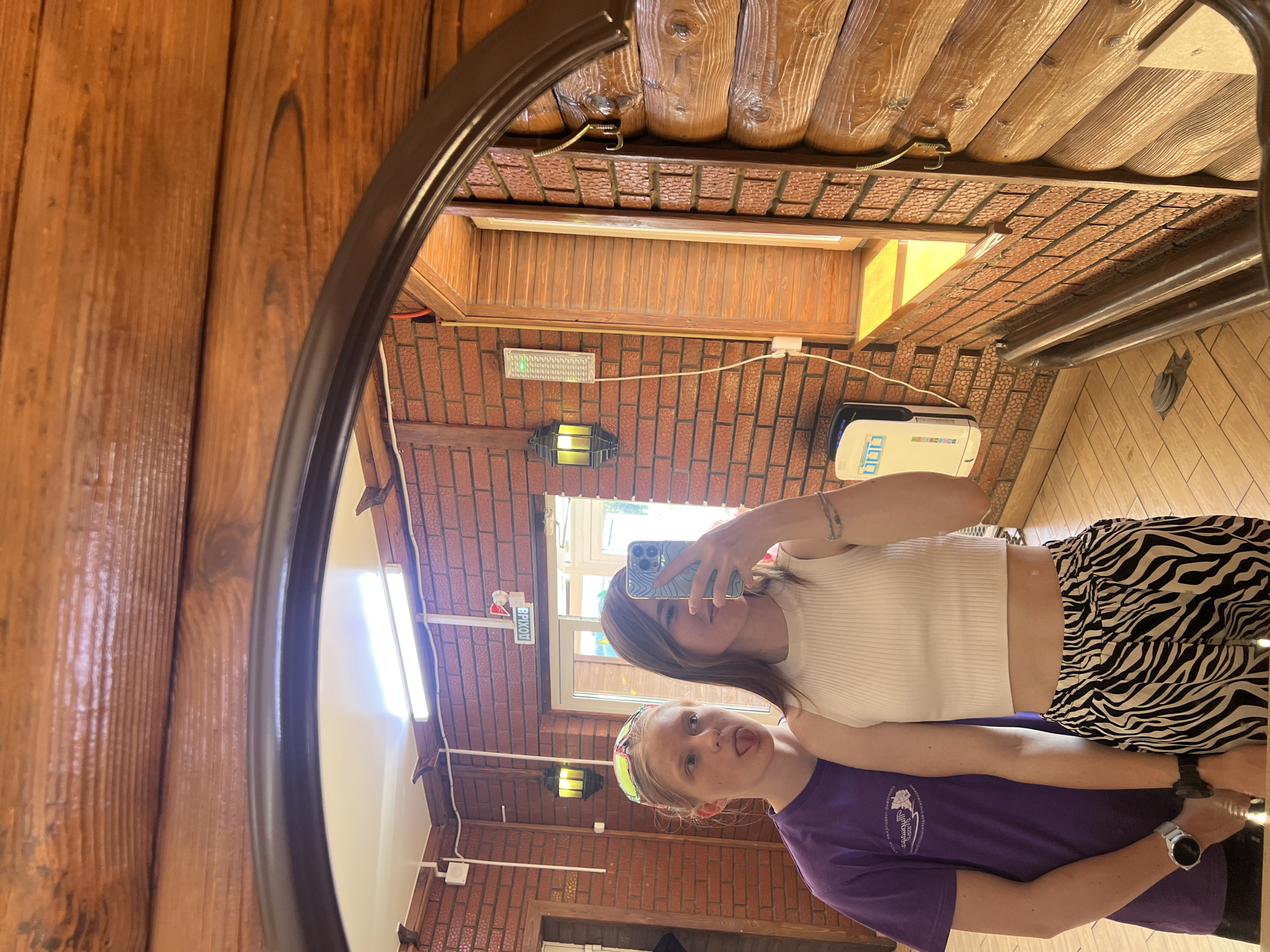Two young women are reflected in a round mirror with a dark brown frame. The woman in the foreground wears a purple t-shirt and a brightly colored headband. She is sticking her tongue out. Behind her, the other woman holds a phone, taking a selfie in the mirror. She wears a white tank top and zebra-print pants. The background reflects the interior of a log cabin with brick walls, a wooden ceiling, and fluorescent lighting. A sign with Russian text is visible near a window. The location is possibly in Grishanki, Russia.