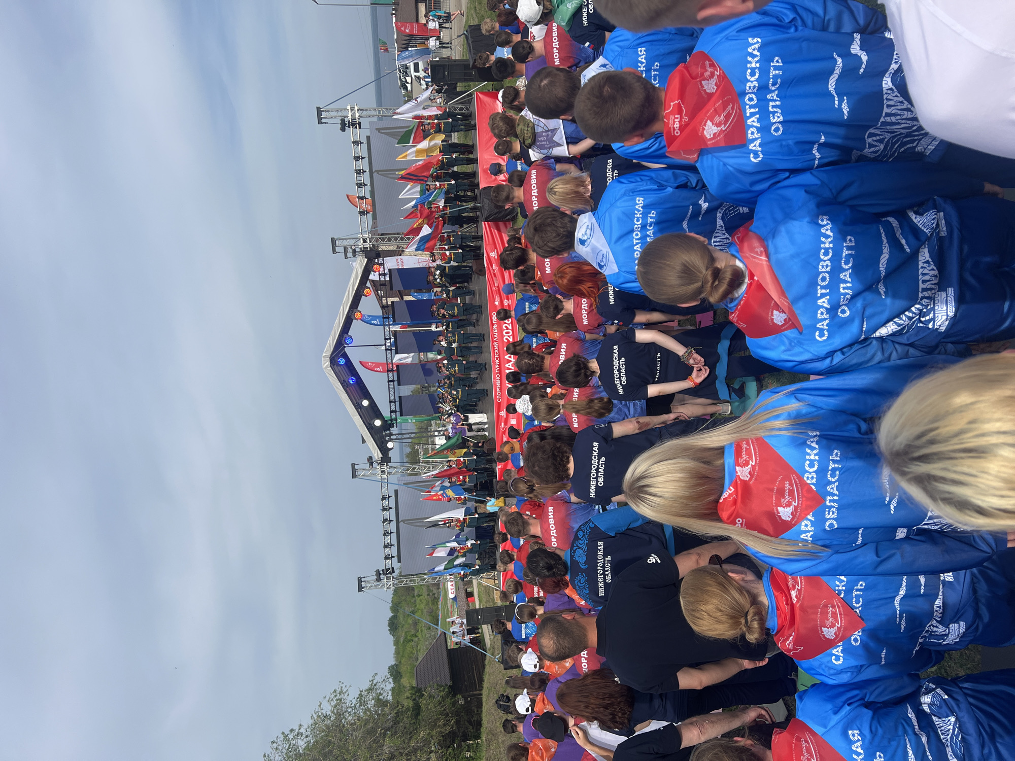 A large outdoor gathering is underway, featuring a stage with flags and event signage. Attendees are seated or standing facing the stage. Many are wearing blue jackets with the text "САРАТОВСКАЯ ОБЛАСТЬ," with some wearing red scarves. The stage area displays flags and a banner promoting "СПОРТИВНО-ТУРИСТСКИЙ ЛАГЕРЬ ПФО 2024," indicating a sports and tourism event. The event location, Khvalynsk, Russia, is implied by the jackets, flags, and event signage. The crowd is multi-aged, and officials in uniform stand on the stage.