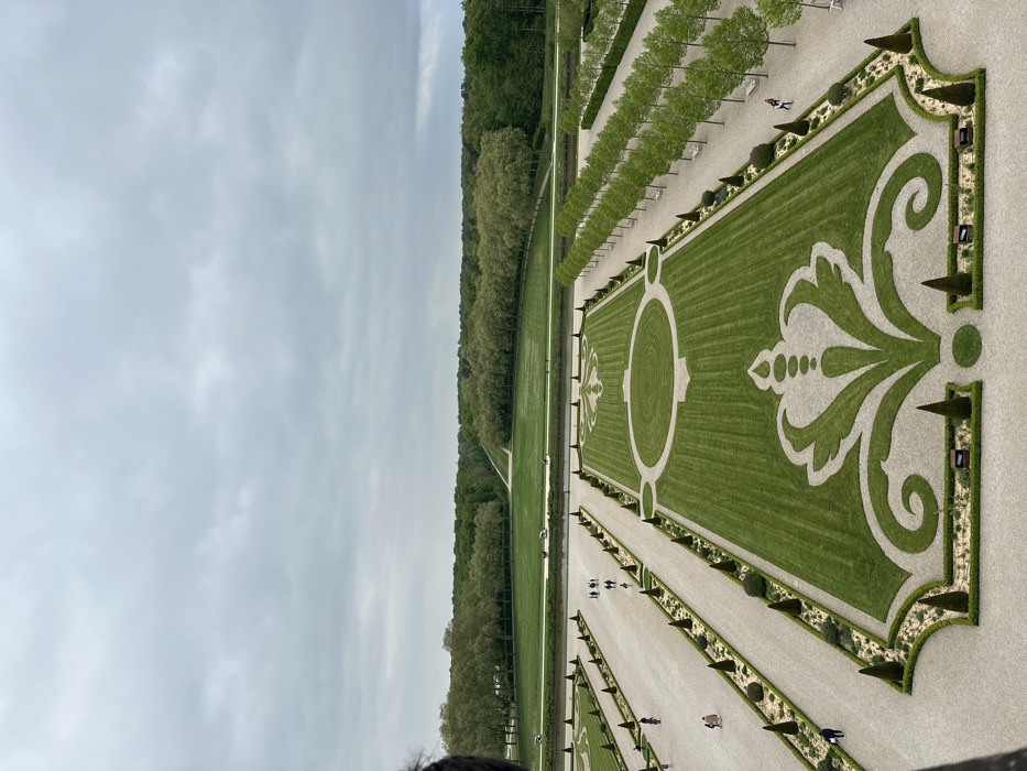 The image shows an aerial view of the formal gardens of the Chateau de Chambord, located in the Loire Valley of France at latitude 47.616, longitude 1.518. A large, manicured lawn with a patterned design extends from the foreground towards a line of trees. The design is outlined with gravel pathways, and there are rows of trees lining the pathways on either side. The background is a wide open field with trees on either side, which appear to be along the banks of a river, before the distant, forested horizon. A few people are walking on the gravel paths, which are lined with small trees and bushes. The sky is overcast with gray clouds. The photograph appears to have been taken from a window on one of the upper levels of the chateau, possibly the roof.