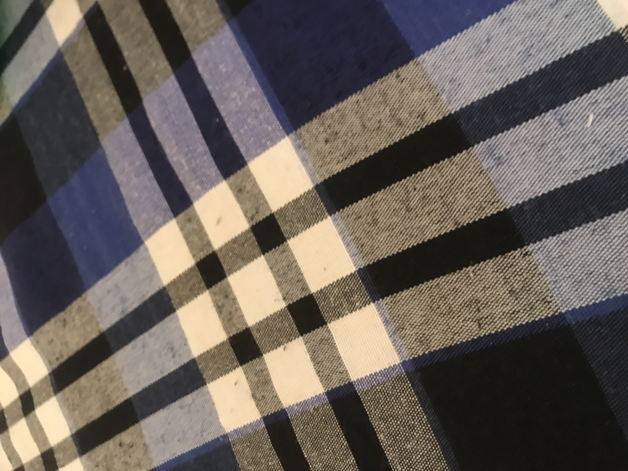 This image displays a close-up, angled view of a plaid fabric surface located in Unknown, Unknown. The fabric features a pattern composed of intersecting bands of dark blue, white, dark grey/black, and a lighter blue. The weave of the fabric is visible, particularly in the white and grey areas, exhibiting a slightly textured, possibly cotton or flannel-like material. Numerous small, dark specks, likely lint or fabric pilling, are scattered across the surface, more noticeable on the lighter-colored bands and in the grey sections. The lighting is soft, highlighting the texture and minor imperfections without casting harsh shadows. No people or specific objects other than the fabric are discernible within the frame.