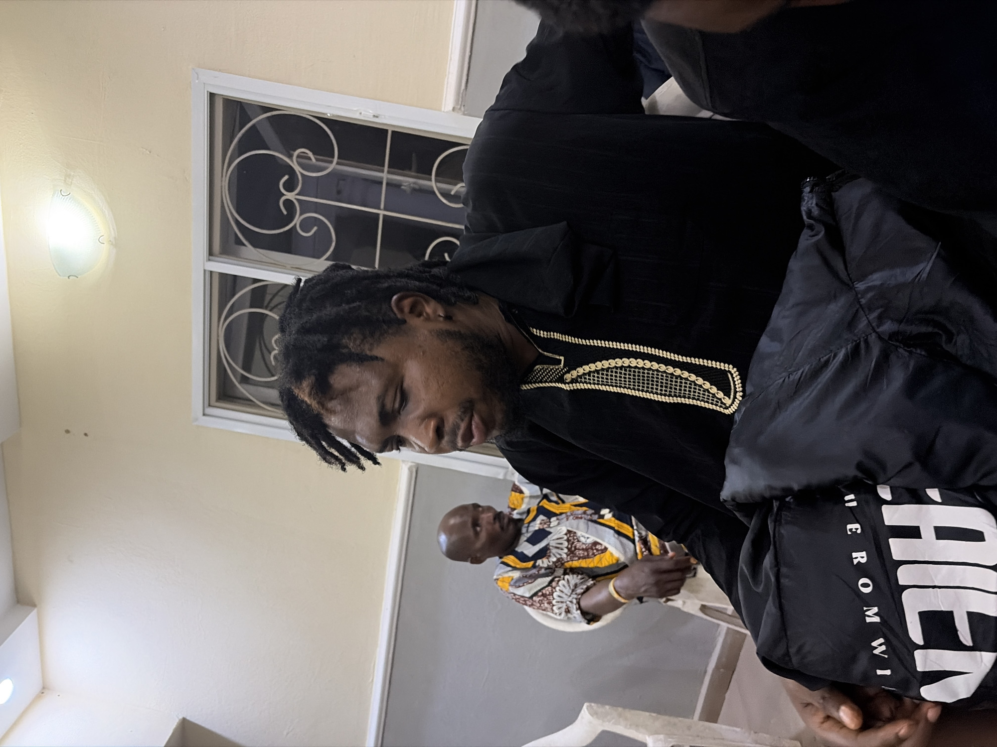 The image depicts an interior scene, possibly in Jos, Nigeria, judging from the clothing worn by the individuals. The main subject is a man with dreadlocks wearing a black, traditional-looking outfit with gold detailing around the collar. Behind him, another man is visible, wearing a patterned white, yellow, and black shirt. Both men are seated. In the background, there is a window with a decorative metal grill. The walls are painted in a light beige color, and there's a light fixture on the wall. The image is taken from a slightly elevated angle, making the subjects appear to be looking downward. The exact location and actions occurring are not clear, but the attire suggests a potentially formal or culturally significant gathering.
