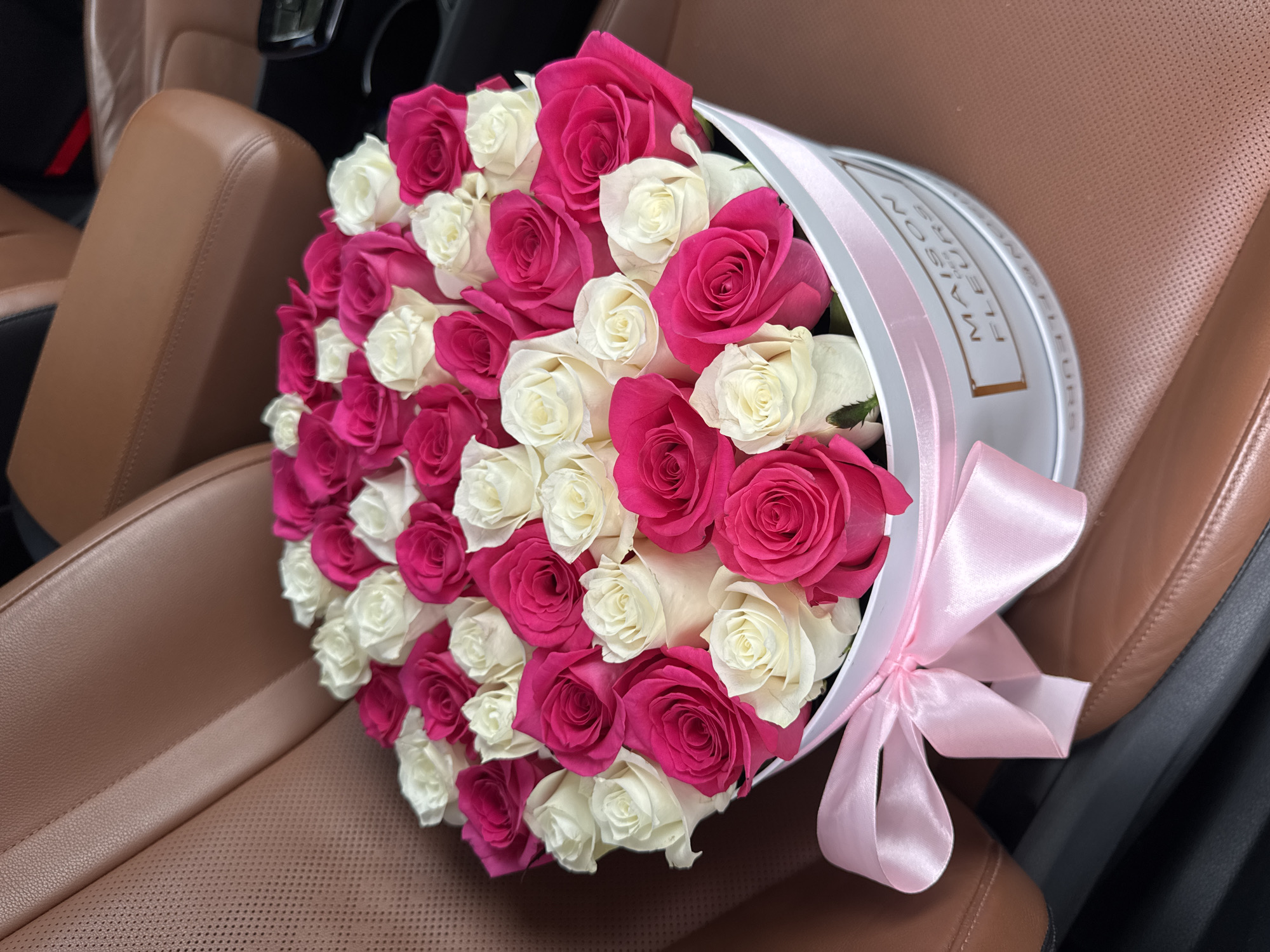 A close-up shot captures a vibrant bouquet of pink and white roses arranged in a round, white hatbox, accented with a pink ribbon tied in a bow. The hatbox is positioned on a plush, brown leather car seat. The textured leather, with its subtle perforations in some areas, suggests a luxurious interior, possibly an SUV or a high-end sedan. The background is blurred, hinting at the inside of a car, with a glimpse of dark upholstery and a seatbelt. The lighting indicates it's daytime, possibly a sunny day given the brightness of the scene. The roses themselves are full and well-formed, with some petals slightly unfurled, showcasing their delicate beauty. The pink roses are a deep, saturated fuchsia, while the white roses offer a soft contrast. The hatbox has text on its side, appearing to read "MAISON FLEURS" in a stylized font. The overall impression is one of a thoughtful gift, presented elegantly within the comfort of a car, hinting at a special occasion or a gesture of affection. The setting suggests the photo was taken in Almaty, Kazakhstan, adding a specific geographical context to the scene.