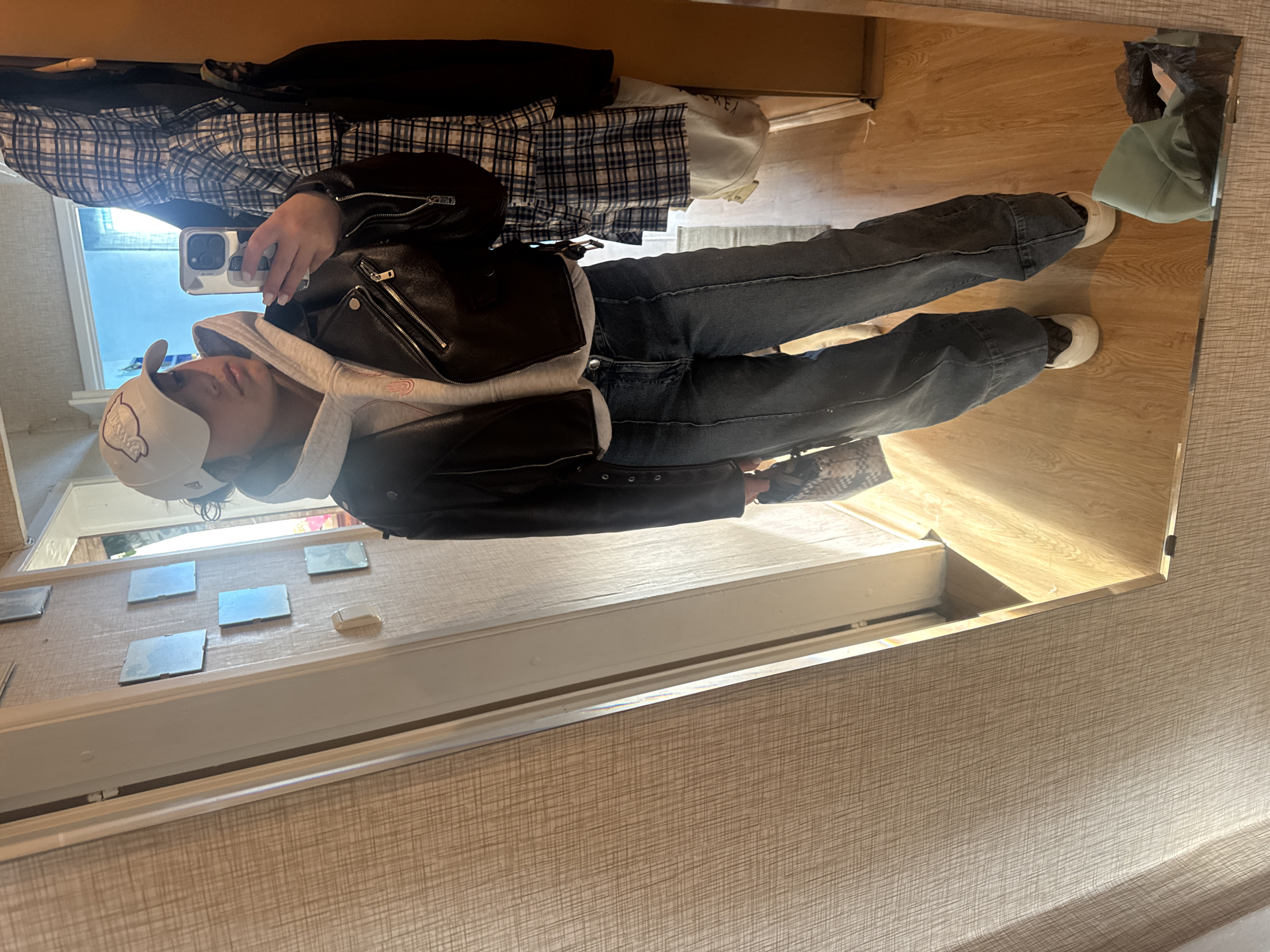 A person is standing in front of a mirror, taking a selfie. They are wearing a white baseball cap, a grey hoodie, a black leather jacket, dark-colored jeans, and white sneakers. In their right hand, they are holding a smartphone. The background shows a wood-grain floor, a wall with small, rectangular mirrors, and various items of clothing on a shelf. The location is Unknown, Unknown.