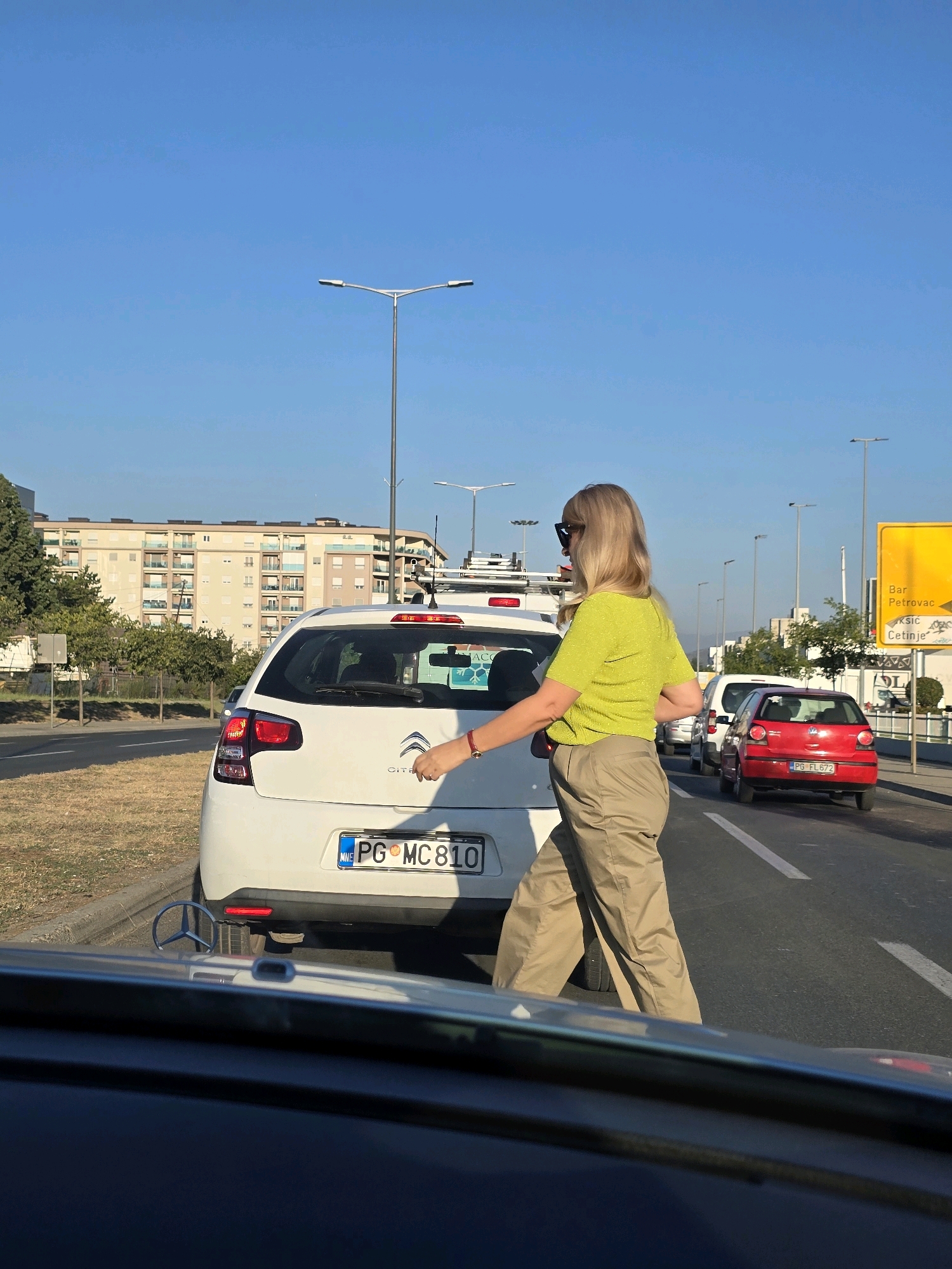 In Podgorica, Montenegro, on a sunny day, a woman with blonde hair and sunglasses walks across a street in moderate traffic. She is wearing a bright yellow top and loose tan pants. She appears to be crossing the street in front of a white Citroen car, which has a license plate displaying "PG MC 810." Other cars, including a red Volkswagen Polo with license plate "PG FL 672," are stopped in traffic behind the Citroen. The background features a multi-story apartment building, trees, streetlights, and a yellow directional sign indicating "Bar Petrovać Cetinje." The perspective is from inside another vehicle, looking forward.