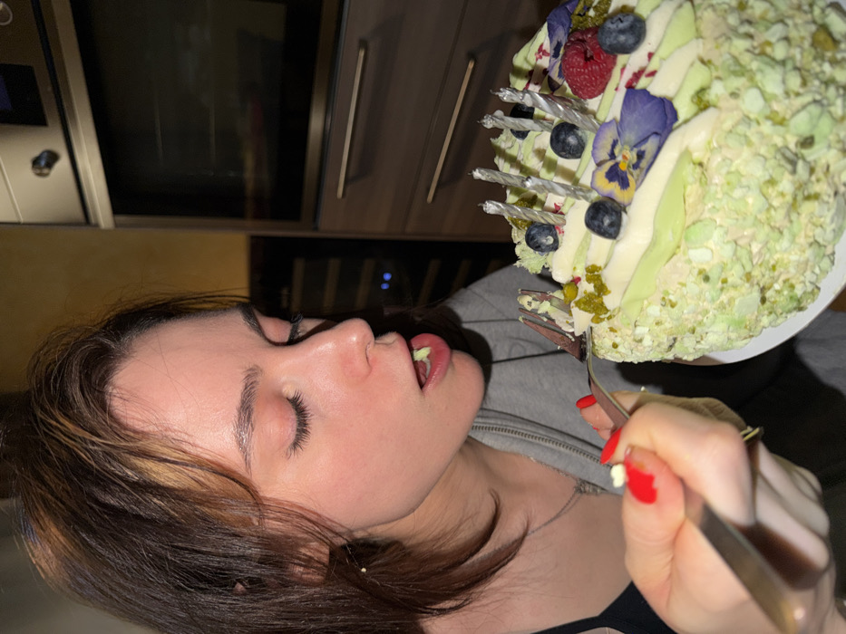 A woman closes her eyes while eating a slice of birthday cake with lighted candles. She has a fork in her hand with a small piece of cake on it. The cake is decorated with blueberries, raspberries, pansies, and green sprinkles. Behind the woman is a kitchen with stainless steel appliances.