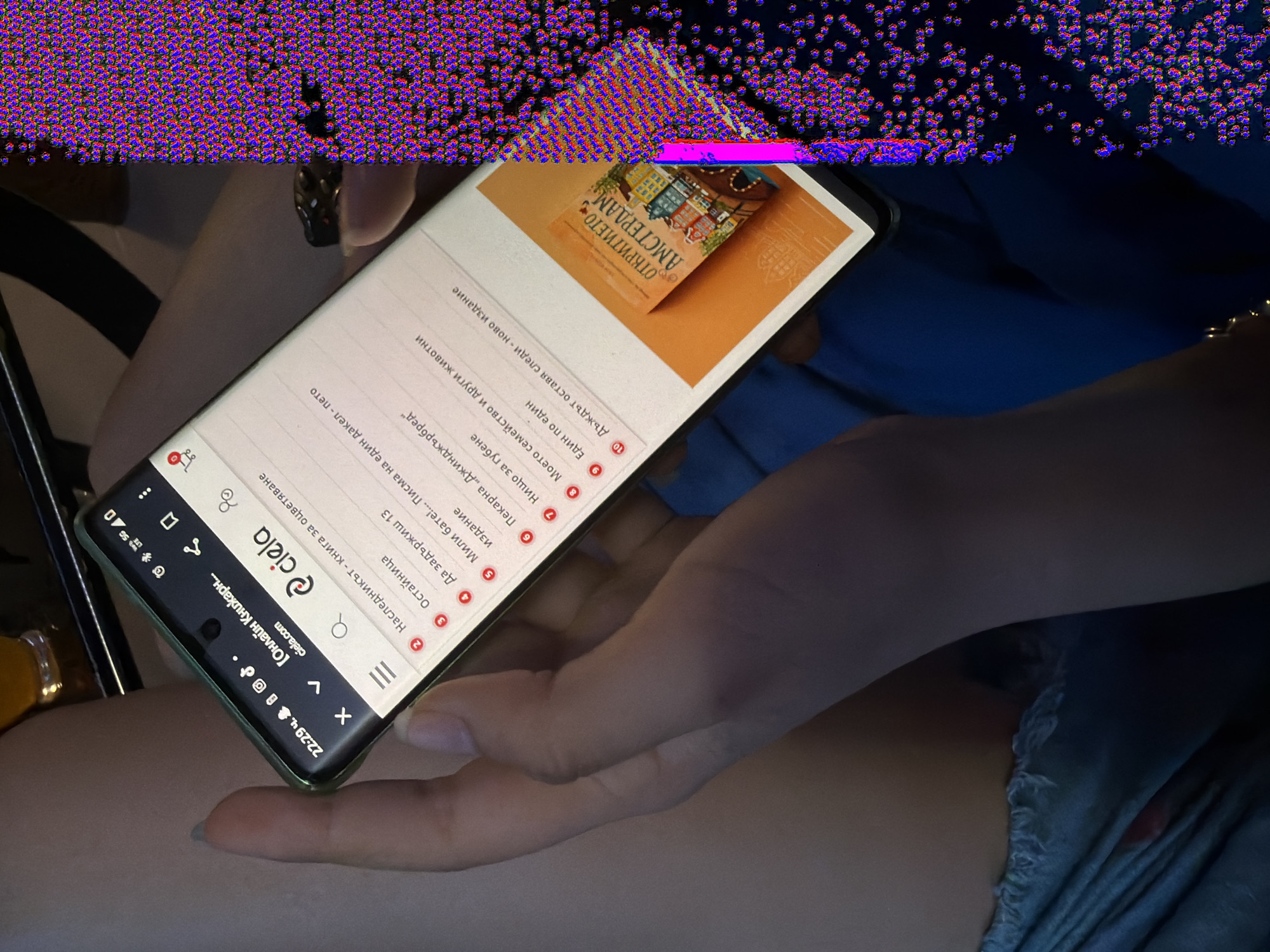 The image shows a close-up of a person's hands holding a smartphone, which displays a webpage from the Bulgarian online bookstore "ciela.com". The webpage lists several books, with the title "Откритието Амстердам" (The Discovery Amsterdam) visible on a book cover in the lower right portion of the phone screen. The person appears to be indoors, possibly in a cafe or at home, with a blurred background of what looks like a table and drink. The overall scene is casual and focused on the smartphone interaction. There is no discernible information about the city of Svilengrad, Bulgaria within the image itself.