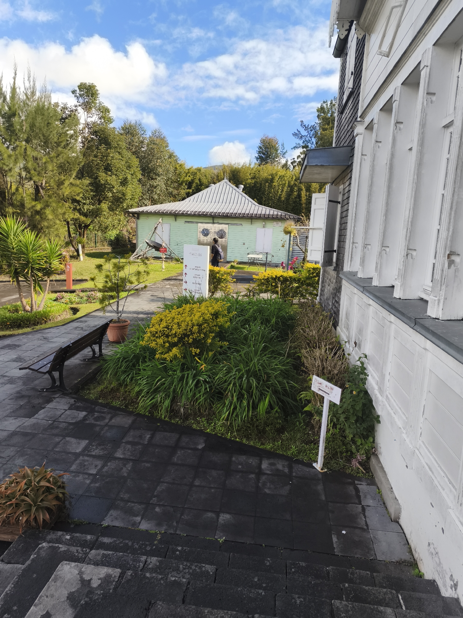 A person is walking away from the camera on a paved path in front of a light green building with a silver roof. The building appears to be a historical structure. To the right of the green building is a tall, white, multi-story building with visible shutters on its windows. There is a wooden bench on the paved path, and various green plants and yellow flowers are in the foreground and around the buildings. The sky is blue with scattered white clouds. There is a sign with arrows and text in front of the yellow flowers. The scene is outdoors and appears to be in a well-maintained park or historical site.