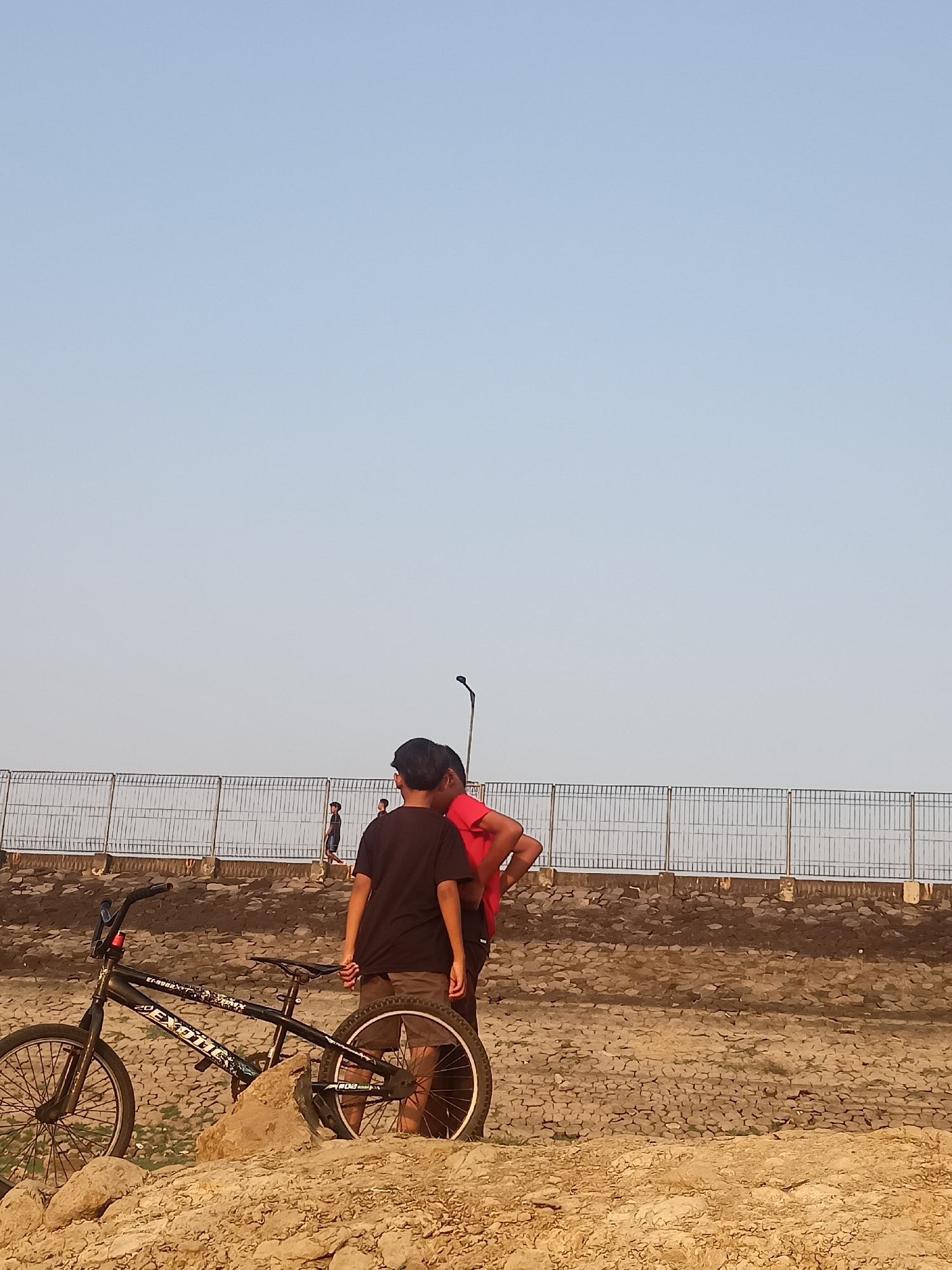 This is a medium shot photograph taken outdoors during the day, likely in Tanon, Indonesia. The sky is a clear, pale blue, taking up the upper two-thirds of the frame. Below the sky, a light-colored metal fence runs horizontally across the middle ground, with a textured, dry ground surface composed of small stones and dirt beneath it. In the foreground, there is a pile of rough, tan-colored rocks and sand.

Dominating the left side of the foreground is a black BMX bicycle. It is positioned on its side, with the front wheel pointing towards the left edge of the frame and the rear wheel towards the right. The bicycle has a sticker on its frame with the word "EXOTIC" prominently displayed.

In the middle ground, to the right of the bicycle, two young males are standing. The male on the left is wearing a black t-shirt and brown shorts. The male on the right is wearing a red t-shirt and dark shorts. They are facing away from the camera, with their backs to the viewer, and appear to be in conversation. A third, smaller male figure is visible further in the background, walking along the path behind the fence. A dark pole with a light fixture at the top is also visible behind the two young males. The overall environment appears to be an outdoor recreational area, possibly near a riverbank or park, given the fencing and the natural ground cover.