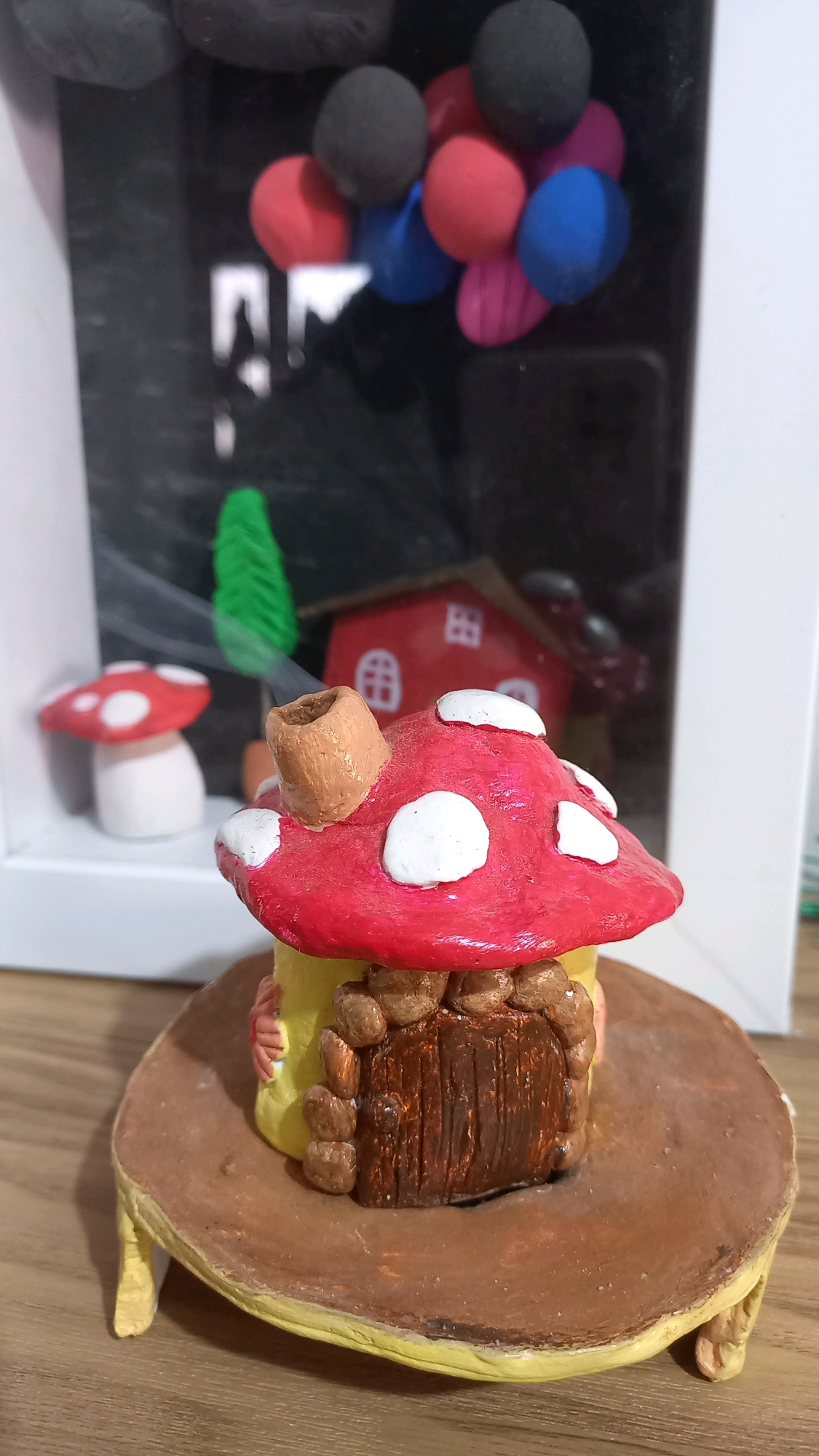 The image shows a close-up, eye-level, outdoor shot of a handmade miniature mushroom house. The mushroom house is red with white polka dots on its cap, and has a yellow cylindrical body with a brown, wooden door surrounded by brown stones. A brown chimney extends from the top of the mushroom cap. The mushroom house sits on a brown, saucer-like base with yellow legs. In the background, there is a miniature red house with white windows, a green miniature tree, and some colorful balloons. The scene is set on a wooden surface. This image does not depict the city of Pammanu, Indonesia.
