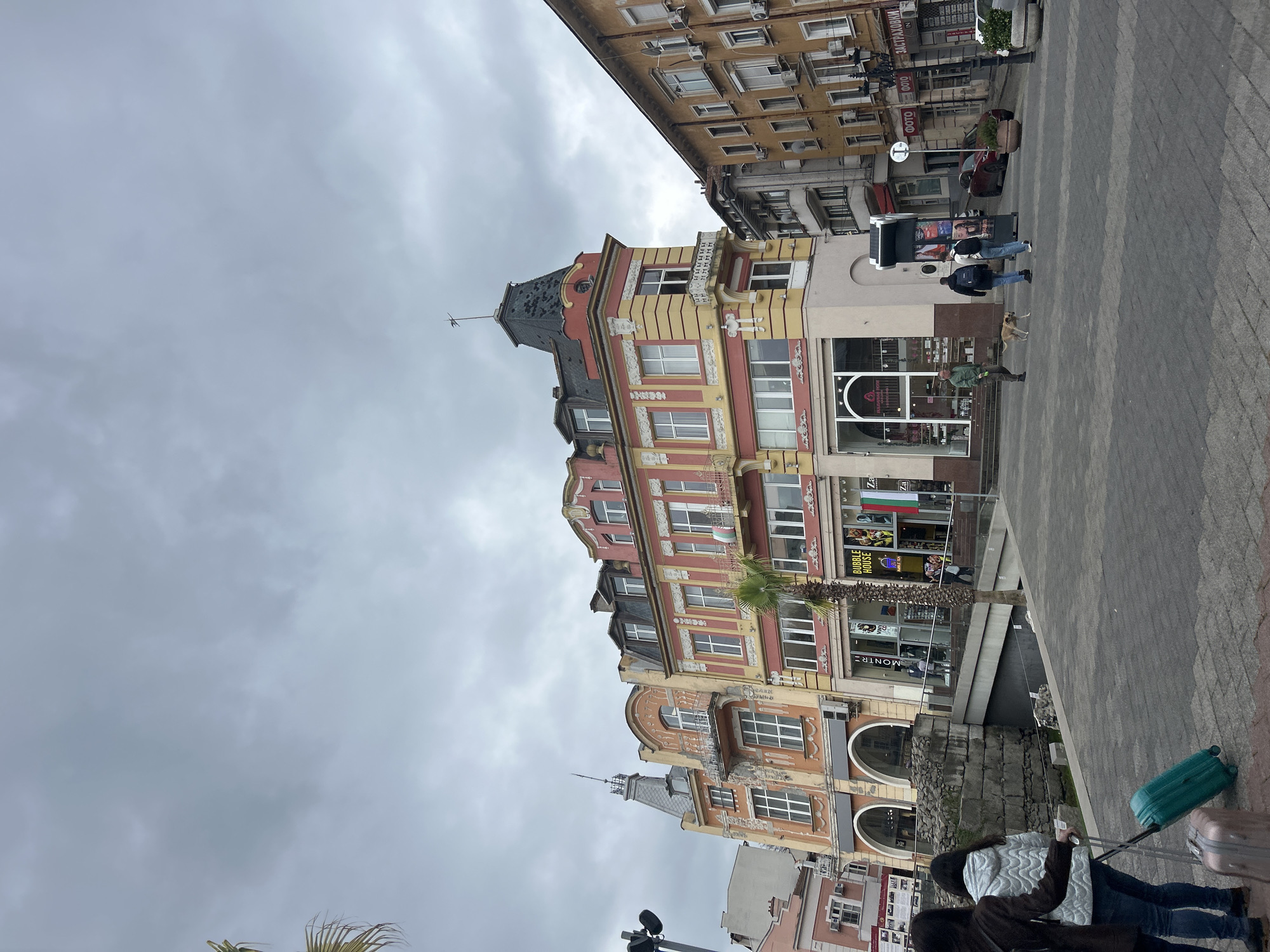 The image depicts a lively street scene in Plovdiv, Bulgaria, under an overcast, grey sky. The central focus is a prominent, ornate multi-story building featuring a distinctive pointed, dark-tiled roof and facades painted in warm shades of yellow, red, and off-white, adorned with decorative architectural details and numerous windows and balconies. Surrounding it are other buildings, including a taller yellow structure with many windows and visible air conditioning units.
On the block-paved street, several people are present. In the foreground, two individuals, one wearing a light quilted jacket and another a dark jacket, are walking away from the viewer, each pulling a piece of luggage – one turquoise suitcase and one pinkish-beige. Ancient stone ruins are visible in the lower left, integrated into the urban environment. Further along, a person in a green jacket stands with a light-colored dog on a leash. Near a black street information display, two other individuals are standing. A dark car is parked on the street in the distance.
A palm tree stands near the central building, adding a natural element. Visible text on signs includes "ЗАСТРАХОВКИ" (Insurance) and "ФОТО" (Photo) on an upper building. On a storefront at the base of the ornate building, "BUBBLE HOUSE BUBBLE TEA" and "MONTR" are legible. A flag displaying the green, white, and red stripes of Bulgaria is also visible. The scene captures a blend of historical architecture and modern urban activity on what appears to be a cool, cloudy day.