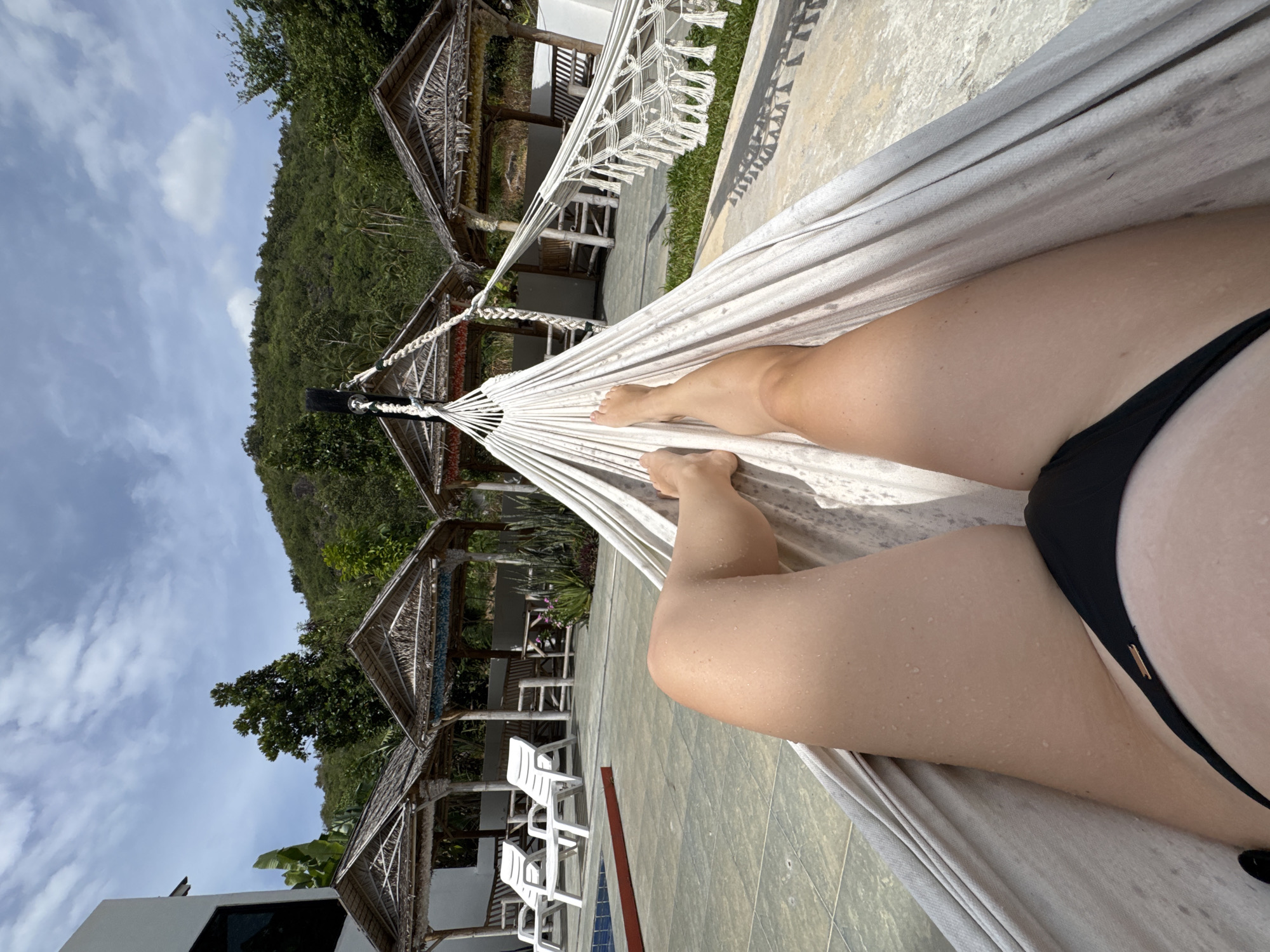 This media file is a first-person perspective photo taken in Baan Taling Ngam, Thailand. The subject is lying in a white hammock, with their legs and part of their torso visible. They are wearing a black bikini. The scene is outdoors, with a lush, green hillside in the background, suggesting a tropical environment. In the middle ground, there are several open-air structures with thatched roofs, possibly bungalows or common areas, and white lounge chairs. The ground appears to be tiled, and there's a glimpse of a swimming pool. The sky is blue with scattered white clouds, indicating it's daytime and likely sunny. The person's skin appears to have water droplets, suggesting they may have just been in the water or it's humid. The overall mood is relaxed and leisurely, consistent with a vacation or resort setting. There is no visible text in the image.