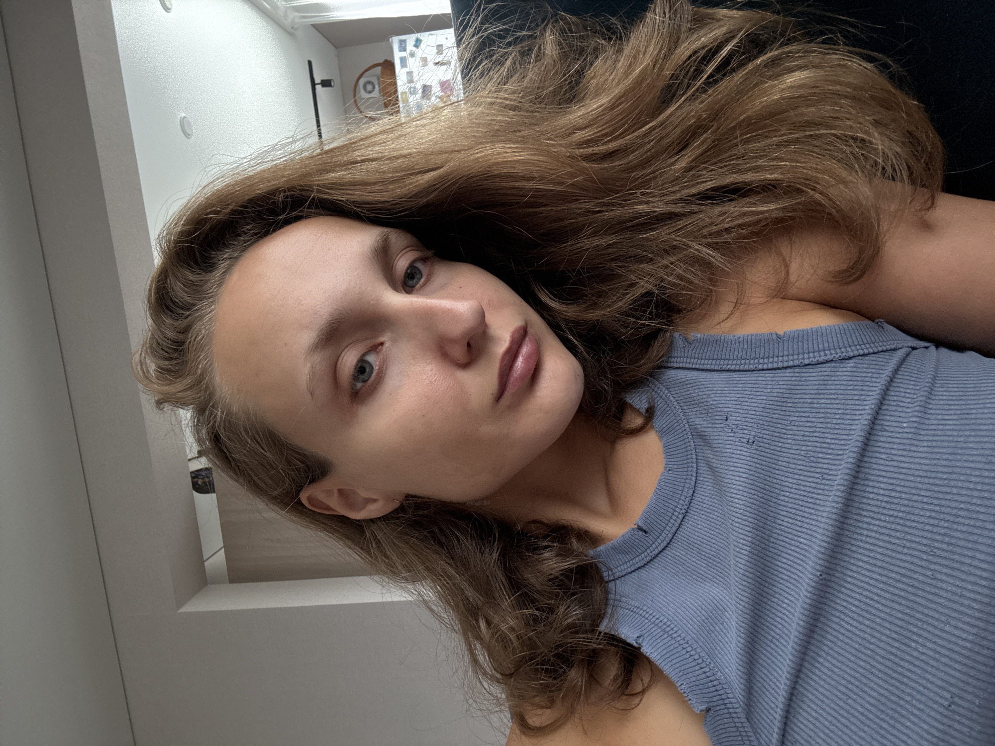 A close-up, head-and-shoulders selfie shows a young woman with long, wavy brown hair and blue eyes. She is wearing a light blue, ribbed tank top with distressed detailing at the straps and neckline. Her skin appears to be bare, with no visible makeup. The background is out of focus but suggests an indoor setting with a white ceiling and wall. The lighting is natural and soft, illuminating her face from the side.