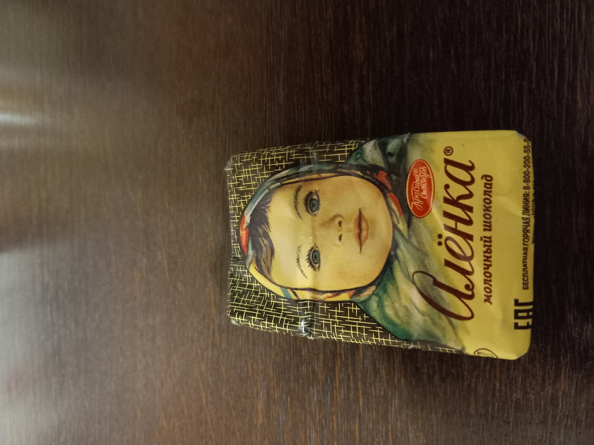 The image shows a chocolate bar wrapper with a traditional design. The wrapper features a drawing of a young girl's face, with blue eyes and rosy cheeks, wearing a headscarf. The background of the wrapper has a patterned design. Visible text on the wrapper includes "Алёнка", which is the brand name, and "МОЛОЧНЫЙ ШОКОЛАД", indicating the product type. There is also a red circular emblem with text inside, and "БЕСПЛАТНАЯ ГОРЯЧАЯ ЛИНИЯ: 8-800-200-55-..." which appears to be a customer service hotline number, though it's partially obscured. The chocolate bar is placed on a dark wooden surface. The lighting suggests it might be indoors, possibly in daylight, though no specific time of day is discernible. The overall impression is of a vintage or classic product.