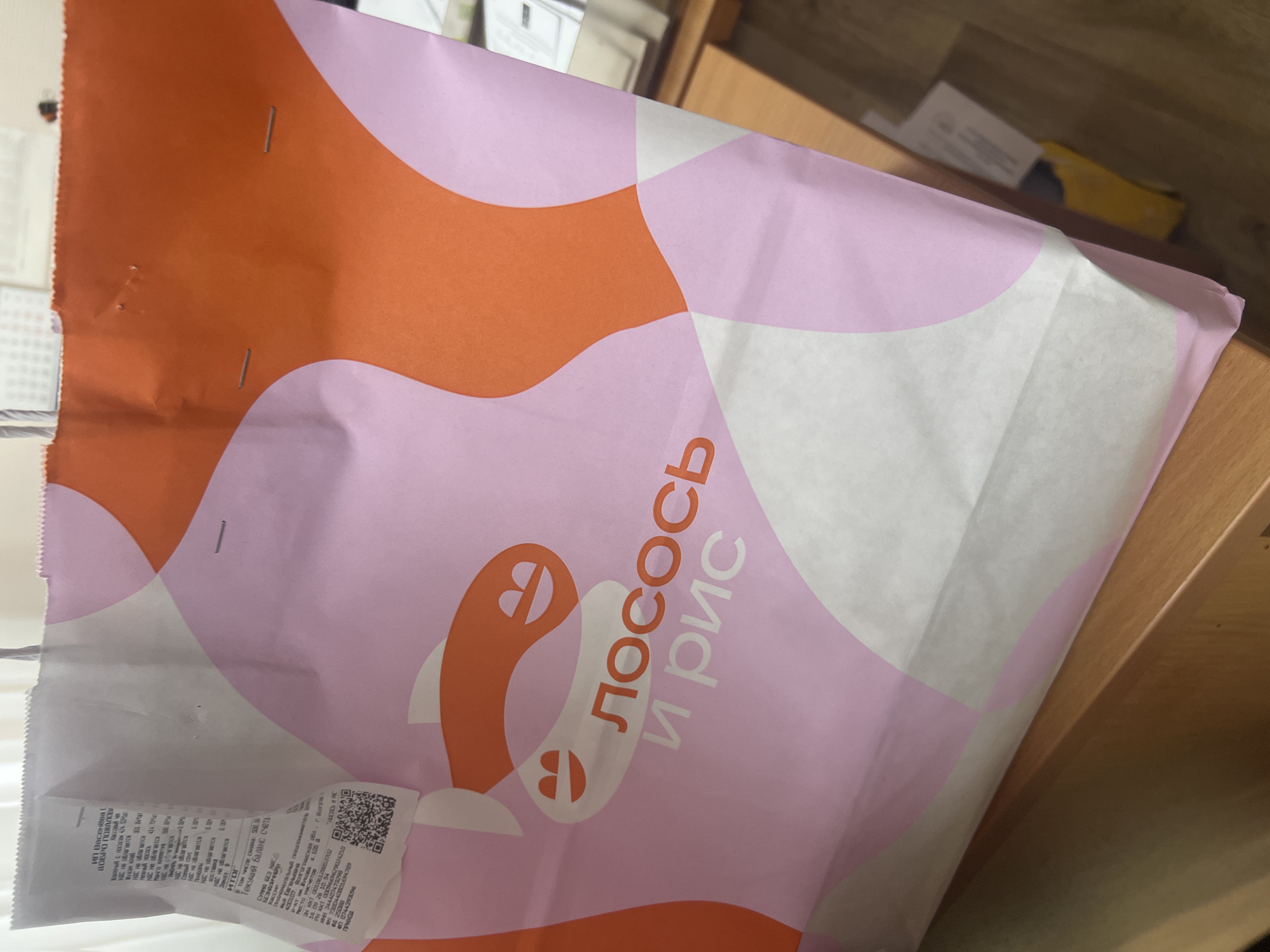 The image shows a paper shopping bag with an abstract design of pink and orange shapes. The bag has text printed on it in Russian, which translates to "Лосось и рис" (Salmon and rice). A portion of a receipt is visible attached to the inside of the bag. The bag is placed on a wooden surface, possibly a desk or table. In the background, there are hints of an indoor environment, including what appears to be a wall and a calendar. The receipt provides information about a transaction, including items, prices, and dates, and mentions "г. Волгоград" which indicates the city of Volgograd, Russia.
