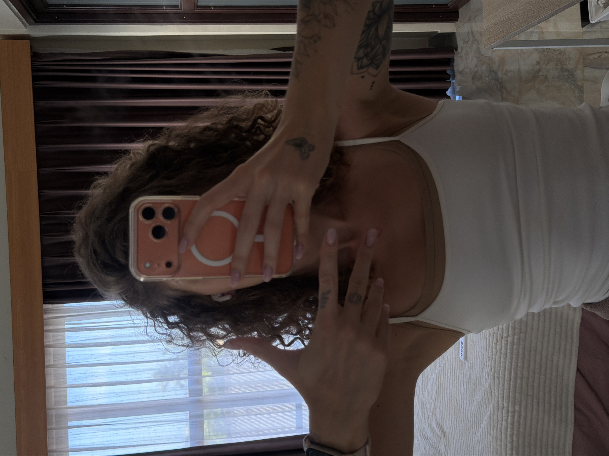 The image shows a person taking a selfie in a bedroom. The person is wearing a white tank top and has long, curly brown hair. They are holding a phone in their right hand, which is reflected in the mirror. There are tattoos visible on the person's arm and hand. The background shows a window with blinds, suggesting it is daytime. The room appears to be a bedroom with curtains and a bed. The overall mood is casual and relaxed. The image is oriented vertically.