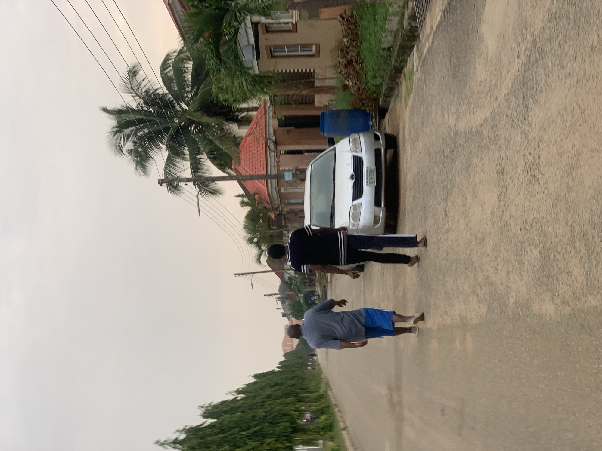 Two men walk away from the camera down a paved road lined with trees and houses in Abuja, Nigeria. The man in the foreground wears a grey short-sleeved shirt and blue shorts. The man behind him wears a black shirt with white stripes on the sleeves and black pants. A silver sedan is parked on the right side of the road. A large blue barrel stands behind the car. The sky is overcast.