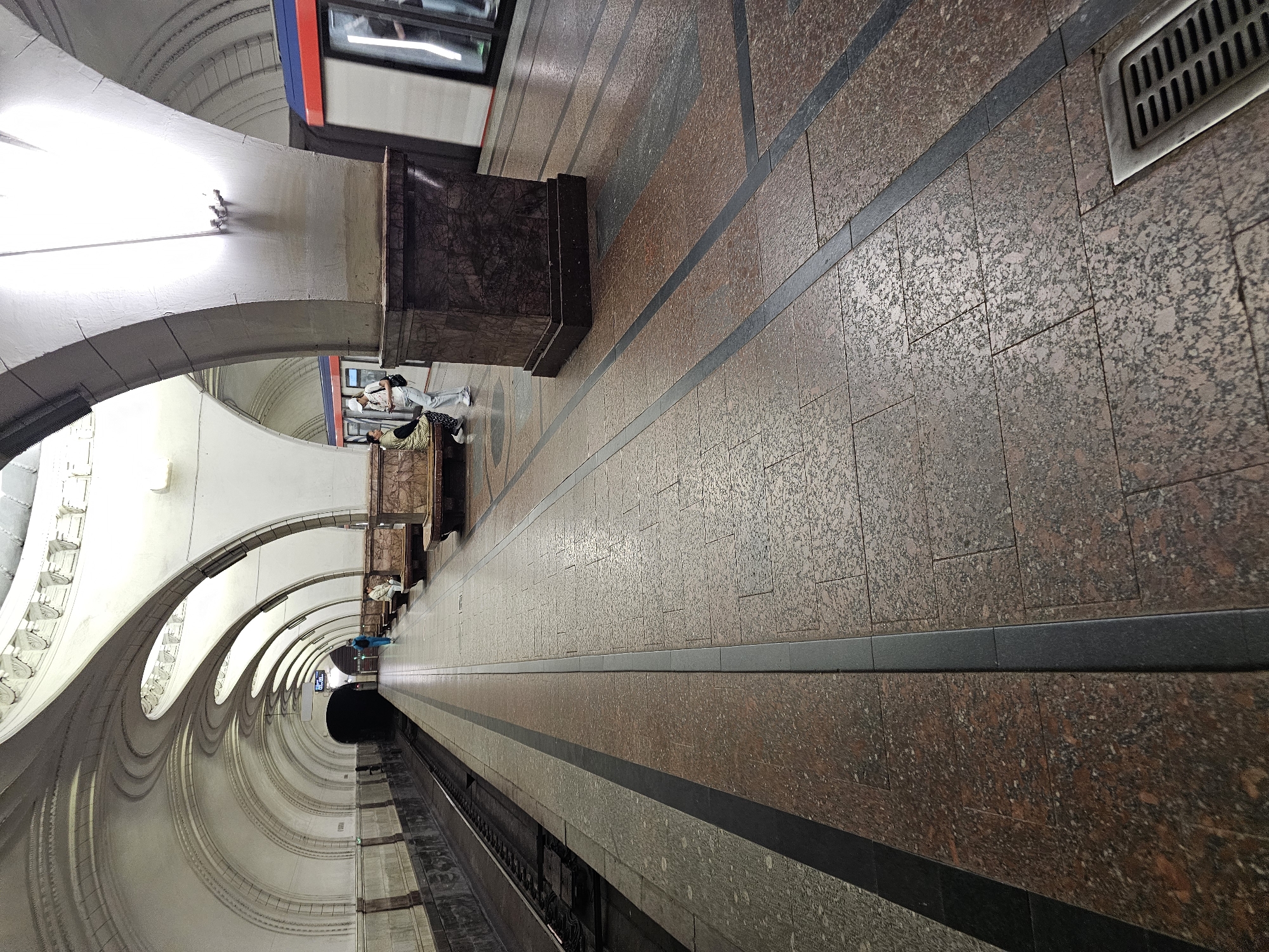 The image depicts an interior view of a metro station, likely in Khimki, Russia, given the presence of a metro train. The station features a grand, arched ceiling with ornate detailing and decorative lighting. The platform is paved with polished granite tiles, accented with dark linear inlays. A train, with red and blue trim, is visible on the right side, partially in motion. Two women are present on the platform; one is seated on a bench, and the other is standing nearby, facing the seated woman. A third person, a woman with a blue backpack, is walking away from the camera towards the darker end of the tunnel. The overall atmosphere is one of a functional and architecturally significant public transit space.