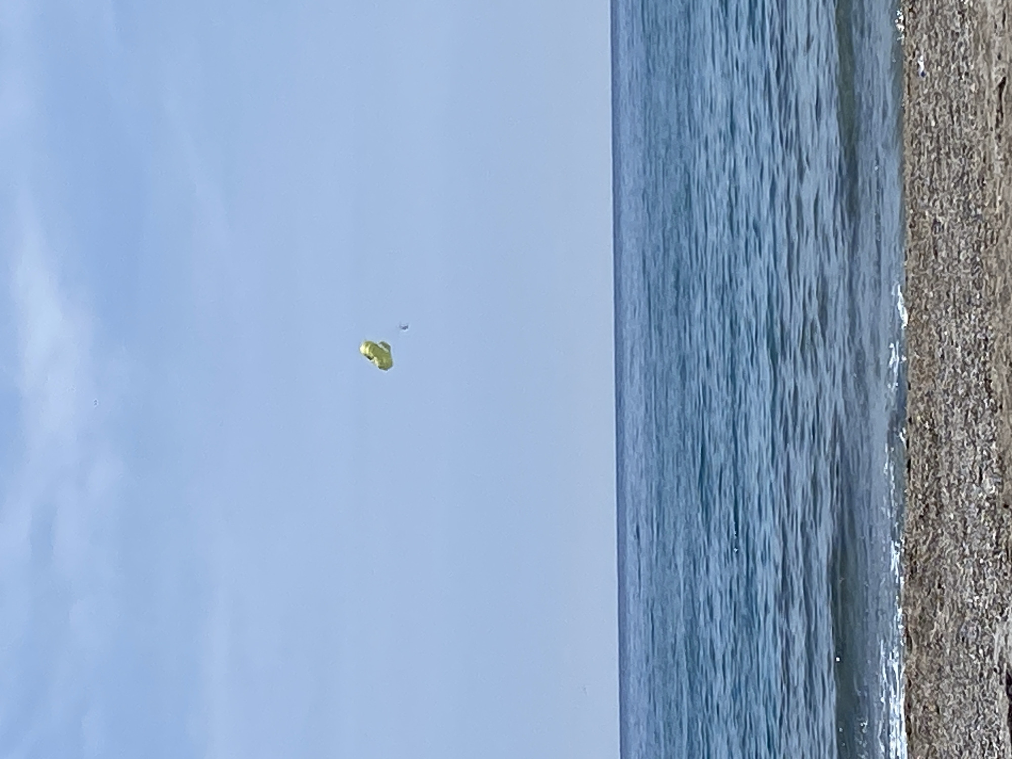 The image depicts a parasailer in the sky above a beach. The parasailer is yellow and appears to be relatively high in the air. Below the parasailer is the ocean, with waves visible along the shore. A sandy beach borders the ocean. The sky is a pale blue. The photograph was likely taken in Alanya, Turkey, given the presence of a parasailer along the beach, a common tourist activity in that city.