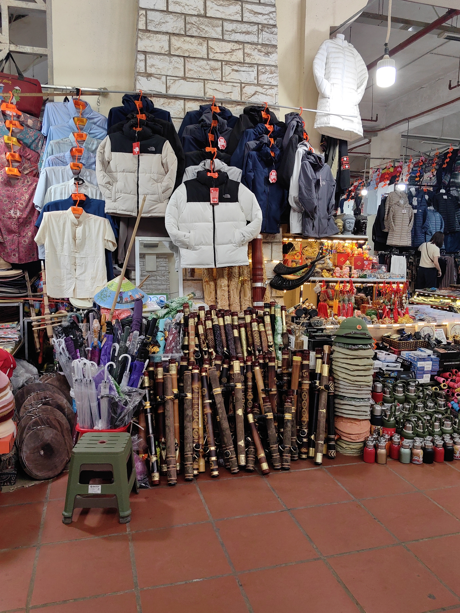 This image captures an indoor market stall in Sa Pa, Vietnam. The floor is composed of reddish-brown tiles. The back wall is light-colored, featuring a section of decorative brick-patterned tiles in the upper left. A bright LED bulb hangs from the ceiling, illuminating the scene.
In the immediate foreground, a dark green plastic stool with a perforated seat is positioned on the tiled floor. To its left, several round wooden cutting boards are stacked vertically, adjacent to a clear plastic bag containing numerous folded umbrellas of various colors. To the right of the stool, a dense display of carved wooden walking sticks or flutes, many with metallic accents, stands upright.
The midground is dominated by merchandise. On the left, multiple shirts and puffer jackets, including several black-and-white two-tone jackets with "The North Face" branding, hang from a metal rail. A long white puffer jacket is displayed on a mannequin torso above this section. Towards the center and right, a multi-tiered display table holds a variety of items: two gray Buddha head statues, small ornate figurines, traditional lanterns, and colorful keychains. A blue Vietnamese conical hat (nón lá) rests near the center. Below this, a stack of khaki-colored folded fabric items is topped by a green military-style cap featuring a red star emblem. Further right, numerous small, colorful thermoses and containers are neatly arranged in rows on the floor and lower shelves.
In the background on the right, one individual is partially visible from behind, standing near a rack of hanging clothing, facing away from the viewer.