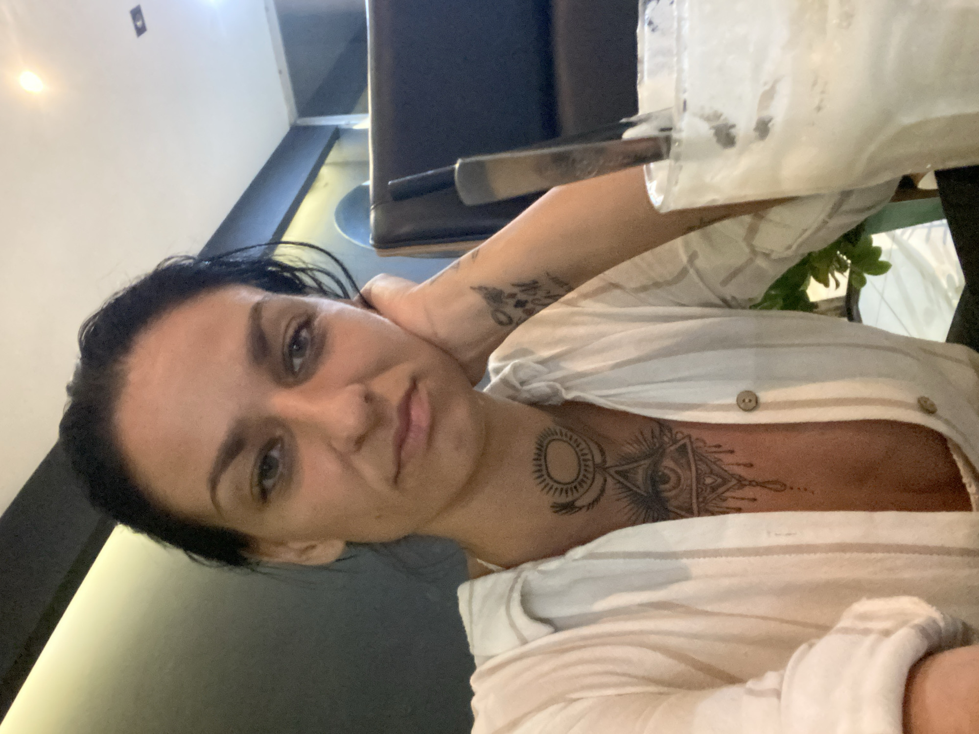 A medium shot depicts a light-skinned woman with dark hair, potentially in Phu Thuy Ward, Vietnam. She wears a partially open, light-colored striped shirt. Tattoos are visible on her left arm and neck/chest. The background features dark furniture and a container partially filled with a white substance.