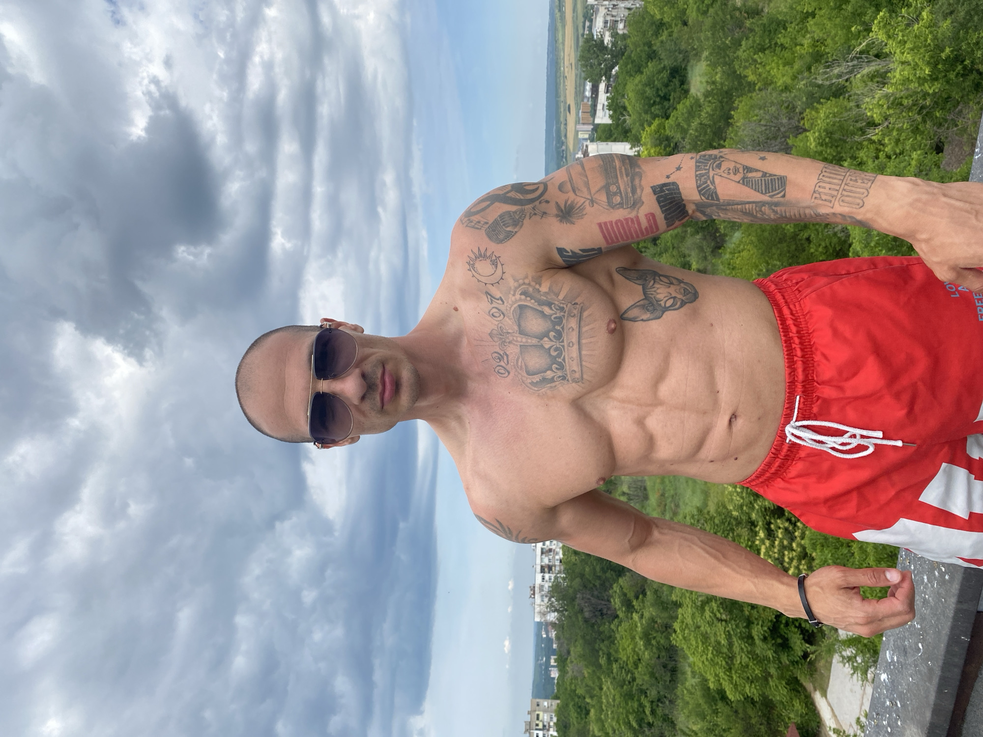 A shirtless man with numerous tattoos on his chest and arms stands outdoors, looking directly at the camera. He is wearing red swim trunks with white accents and sunglasses. The background shows a cityscape with buildings and lush green trees, under a cloudy sky. The location is the city of Pleven, Bulgaria.