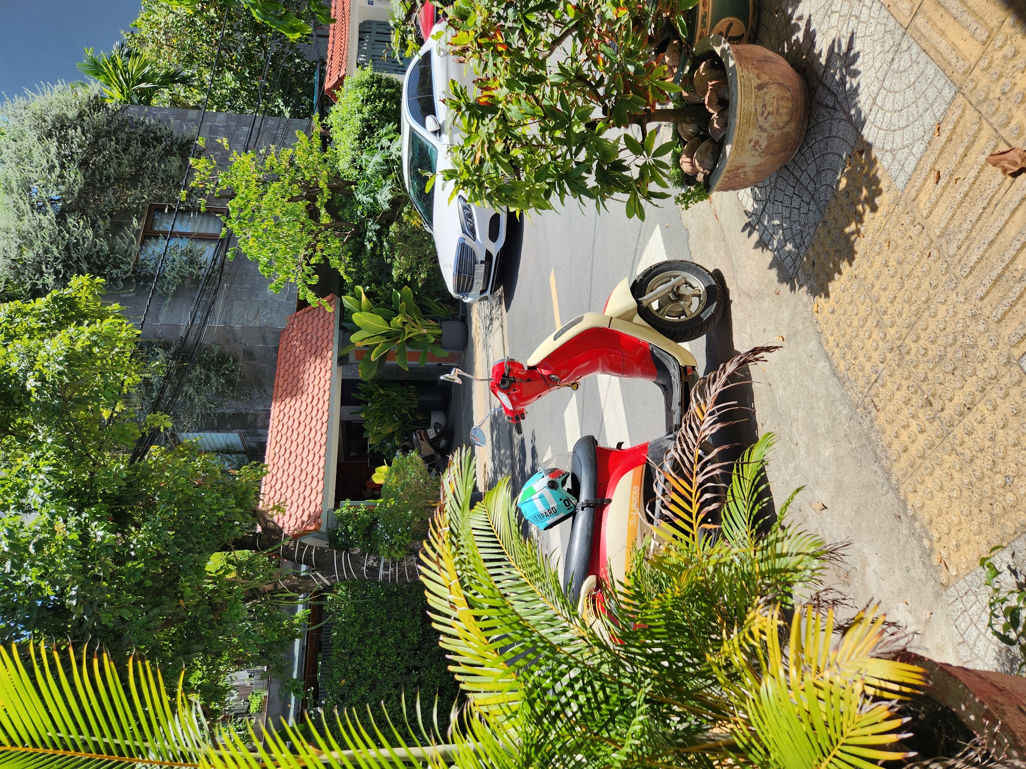 This image depicts a street scene in the city of Da Nang, Vietnam, characterized by a tropical, urban environment. In the foreground, a red and cream-colored scooter is parked on the sidewalk, with a blue and green helmet resting on its seat. Lush green palm fronds frame the left side of the image, casting shadows on the textured yellow tactile paving of the sidewalk. Behind the scooter, a white sedan is parked on the street, displaying a Vietnamese license plate that reads "43A-302.79". The background features buildings with red-tiled roofs and grey stone facades, interspersed with abundant trees and dense vegetation, suggesting a warm, sunny day. The overall scene conveys a sense of everyday life in a vibrant Southeast Asian city.