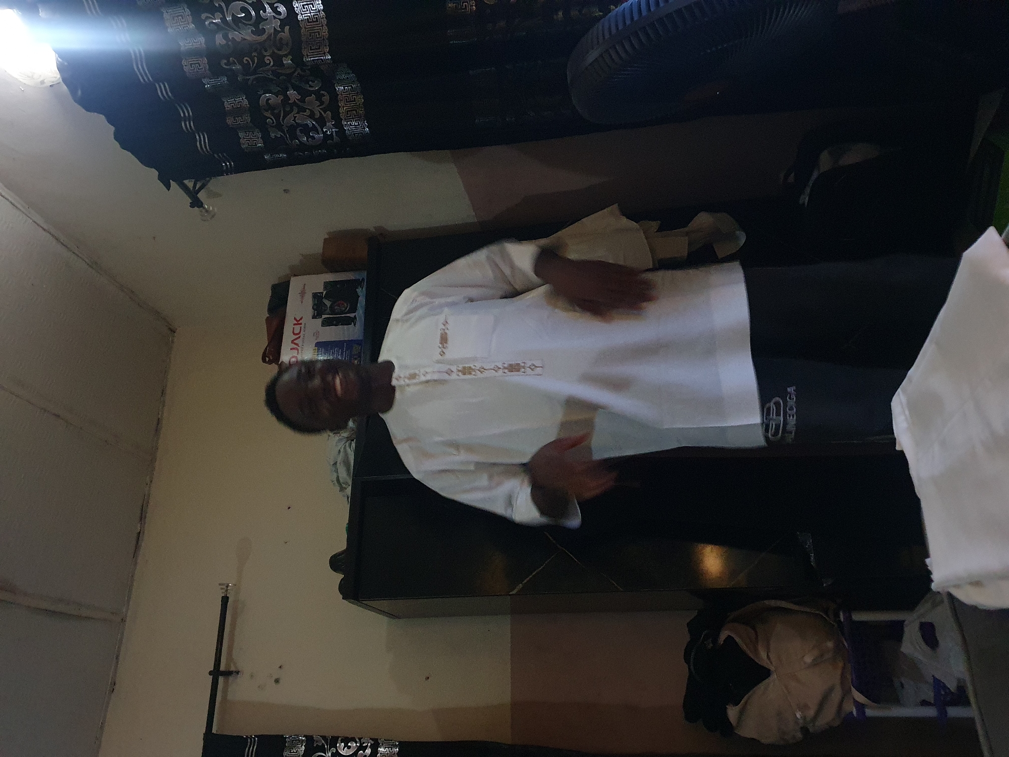 A man is indoors wearing a white shirt and dark pants. He is standing near a dark-colored dresser. There is a box labeled "D Jack" visible near the dresser. The room has a curtain with a patterned design and what appears to be a fan in the upper right corner. The exact location within the city of Lokoja, Nigeria is not determinable from the image.