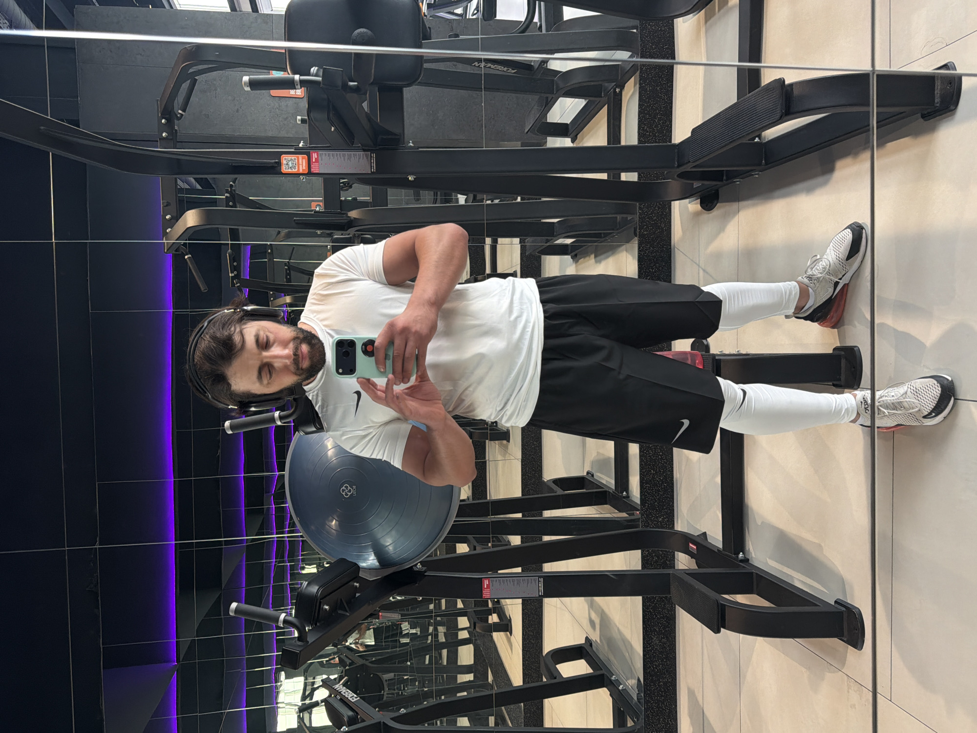 A man is taking a selfie in a gym, likely in the city of Krasnoyarsk, Russia. He is wearing a white T-shirt with a Nike logo, black shorts, white leggings, and sneakers. He has headphones on and is holding a light-green smartphone in his right hand. The gym has black gym equipment with silver accents, including a balance ball, and is mirrored on the left side. The walls are tiled, with black tiles on the left and white tiles on the right. The lighting appears to be a mix of natural and artificial, with purple accents on the mirrored wall.