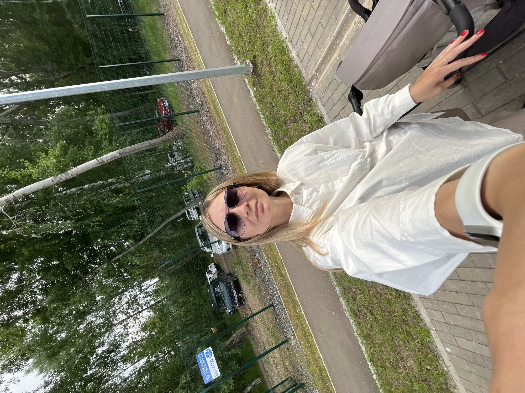 The image shows a blonde woman with sunglasses and a white shirt, taking a selfie outdoors. She is sitting next to a stroller, and her right hand, with red nail polish, is resting on the stroller. The background suggests an outdoor setting with a paved walkway, grass, trees, and a fence. There are cars visible in the distance behind the fence, and a blue sign with white text is also present. The lighting indicates it is daytime. The woman's expression is neutral.
The visible text on the blue sign appears to be in Russian. The words "ВЫХОД" (VYKHOD), meaning "EXIT", and "НА ТЕРРИТОРИИ" (NA TERRITORII), meaning "ON THE TERRITORY" or "IN THE AREA", are legible. There is also a symbol resembling a pedestrian with a leash. The overall context suggests a public area, possibly a park or a recreational facility in Chaikovsky, Russia, with specific signage related to exiting or a designated area.
