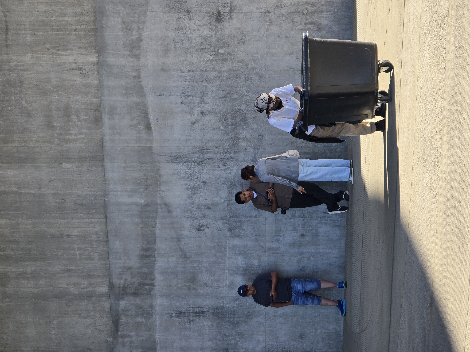 A group of four people stand in front of a large, textured concrete wall. Two men are standing close together on the left, both looking at their phones. One is wearing a blue baseball cap, sunglasses, a dark t-shirt, and denim shorts. The other man is wearing a brown shirt, dark pants, and holding a camera. A woman in a grey jacket and white pants stands to the right of them, facing the two men. Further to the right, a person in a white shirt, khaki pants, and a patterned headscarf is leaning over a large, black wheeled bin. The ground in front of the wall is a smooth, light-colored concrete surface with some circular markings, and a diagonal shadow falls across it from the right. The scene appears to be outdoors in a public space, possibly a park or promenade, in Los Angeles, United States of America.