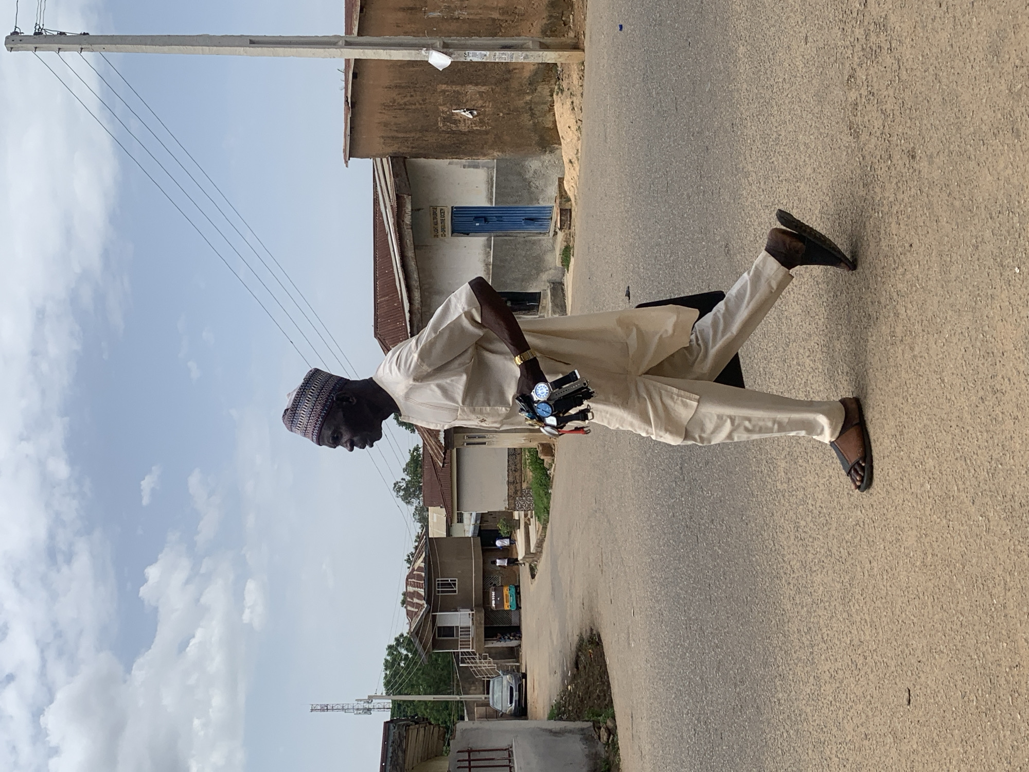 A man is standing on a paved road in Jos, Nigeria. He is wearing a light-colored, traditional outfit and sandals. He has a patterned hat on his head. In his left hand, he holds multiple wristwatches. The background includes buildings of various sizes and styles, a utility pole with overhead lines, and a cloudy sky. A vehicle is also visible in the background.