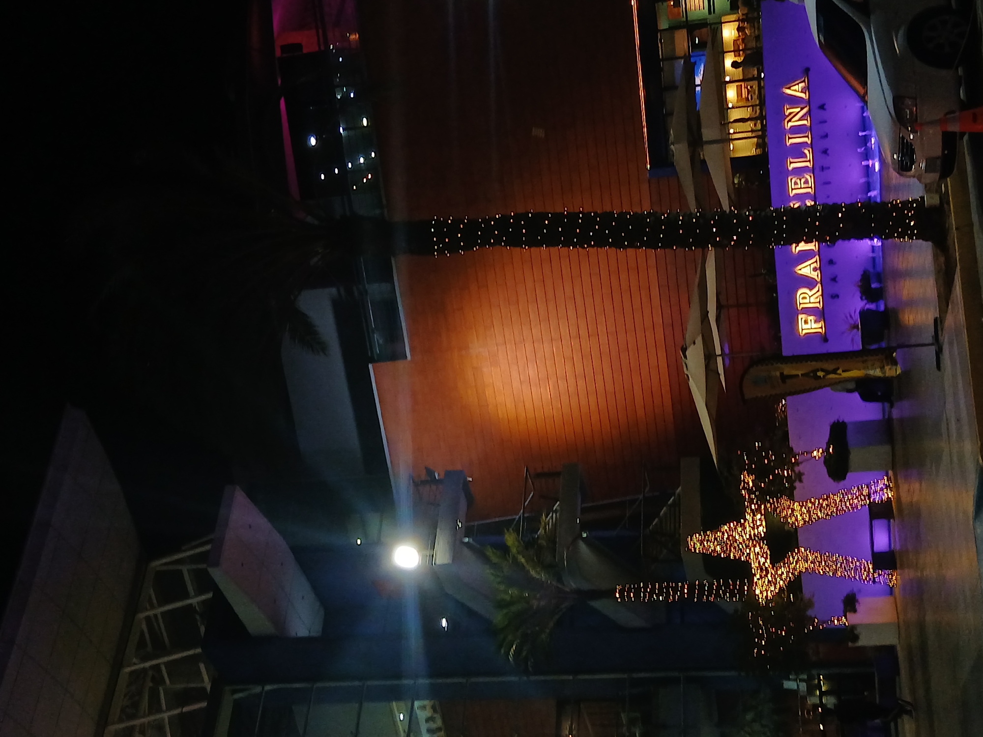 At night in Porlamar, Venezuela, the "Francelina Italia" restaurant is visible. A white car is parked nearby. Illuminated palm trees and decorative lights are prominent. The surrounding architecture suggests a modern commercial area.