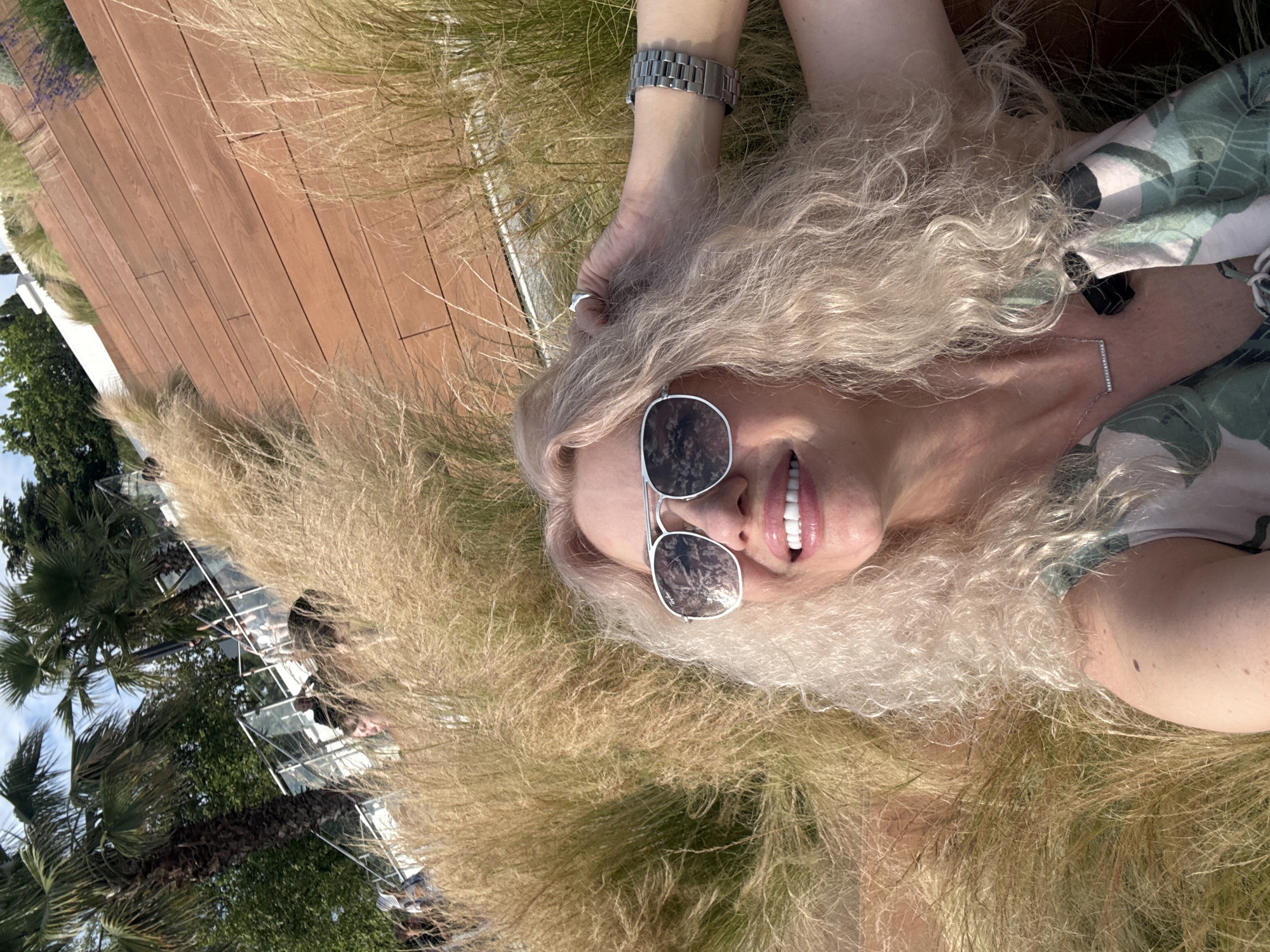 A woman with blonde, curly hair lies on her back in tall, dry grass. She is wearing sunglasses with silver frames and a light-colored top with a green leaf pattern. A silver watch is visible on her left wrist, and a delicate silver necklace with a bar pendant is around her neck. Her mouth is slightly open, revealing white teeth and pink lips, and she appears to be smiling or laughing. The scene is outdoors, with a wooden deck visible to her left and palm trees and a building with glass balconies in the background. The lighting suggests it is daytime, and the dry grass indicates warm weather. The image has a selfie perspective, with the woman looking up towards the camera. The overall emotion conveyed is relaxed and joyful.