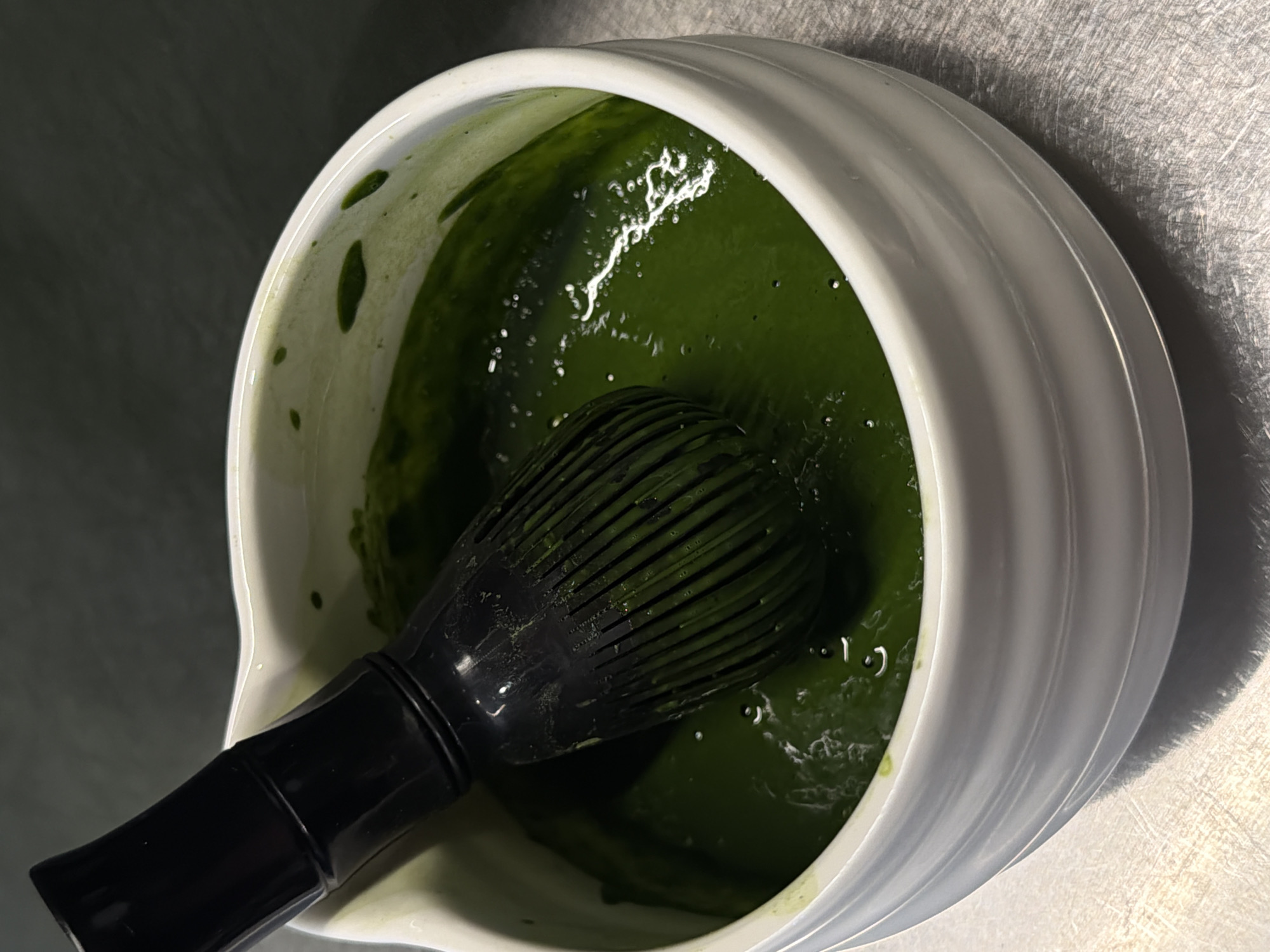 A black bamboo whisk is submerged in a vibrant green liquid within a white, ribbed bowl. The bowl rests on a brushed metal surface. The background is dark and out of focus, suggesting an indoor setting. The image is a close-up shot, with the whisk and bowl occupying the majority of the frame. The green liquid appears to be matcha tea. There are no people or other objects visible in the image. The scene is forensic in its detail, focusing on the texture, color, and arrangement of the objects. The location in Sofia, Bulgaria is not discernible from the image.
