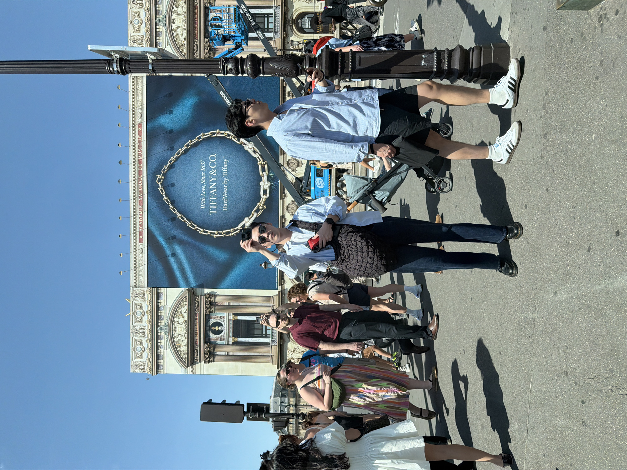 Here is a description of the image.
The image is taken at street level and shows a Tiffany & Co. billboard. The sign appears to be in Paris, France, given the French text "ACADEMIE NATIONALE DE MUSIQUE ET DE CHOREGRAPHIE" which appears at the top of the billboard.
Several pedestrians stand on the sidewalk in front of the billboard. They appear to be tourists, as two individuals have their cameras pointed at the billboard and adjacent building.
A blue SINOBOOM construction lift vehicle is visible above the billboard.