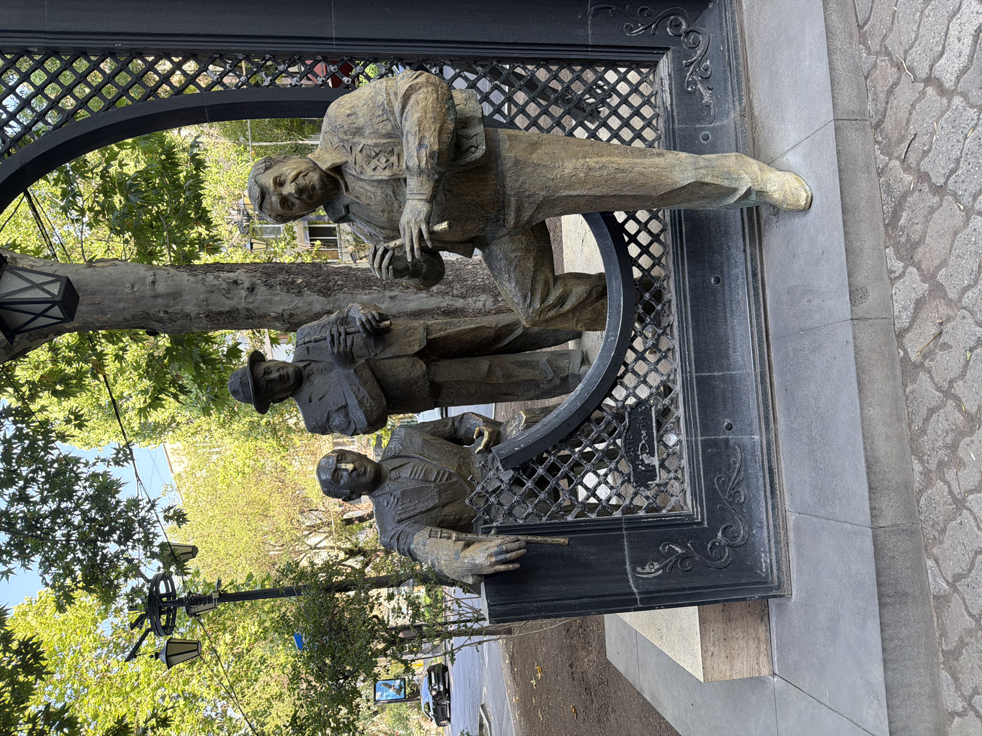 This is a full, slightly high-angle, outdoor shot of a bronze sculpture of three men playing musical instruments in a city square. The men are depicted in formal attire, each holding a flute-like instrument. The sculpture is set against a black metal archway with a diamond-shaped lattice pattern. The man on the right, who is closer to the viewer, is standing with one leg bent and leaning on the archway. The man in the middle is standing and looking towards the right. The man on the left is seated and leaning on a black stone base. Behind the sculpture, trees with green leaves and a streetlight are visible. The ground in front of the sculpture is paved with grey stones and cobblestones. In the background, a street with a black car and a building can be seen. The lighting suggests it is daytime, with sunlight filtering through the trees. The sculpture appears to be a monument, possibly commemorating musicians.