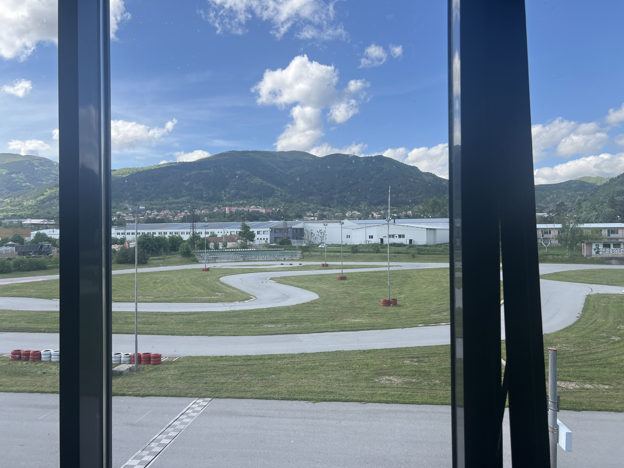 The image shows a go-kart track viewed through a window. The track is paved and winds through a grassy area. Red and white tire barriers are positioned along the track's edges. A mountain rises in the background under a partly cloudy sky. Buildings are visible between the track and the mountain. The location is likely the go-kart track in Tvarditsa, Bulgaria, based on the landscape and local features. No people are visible. The window frame is dark and divides the view. A checkered pattern marking, is visible at the bottom of the window.