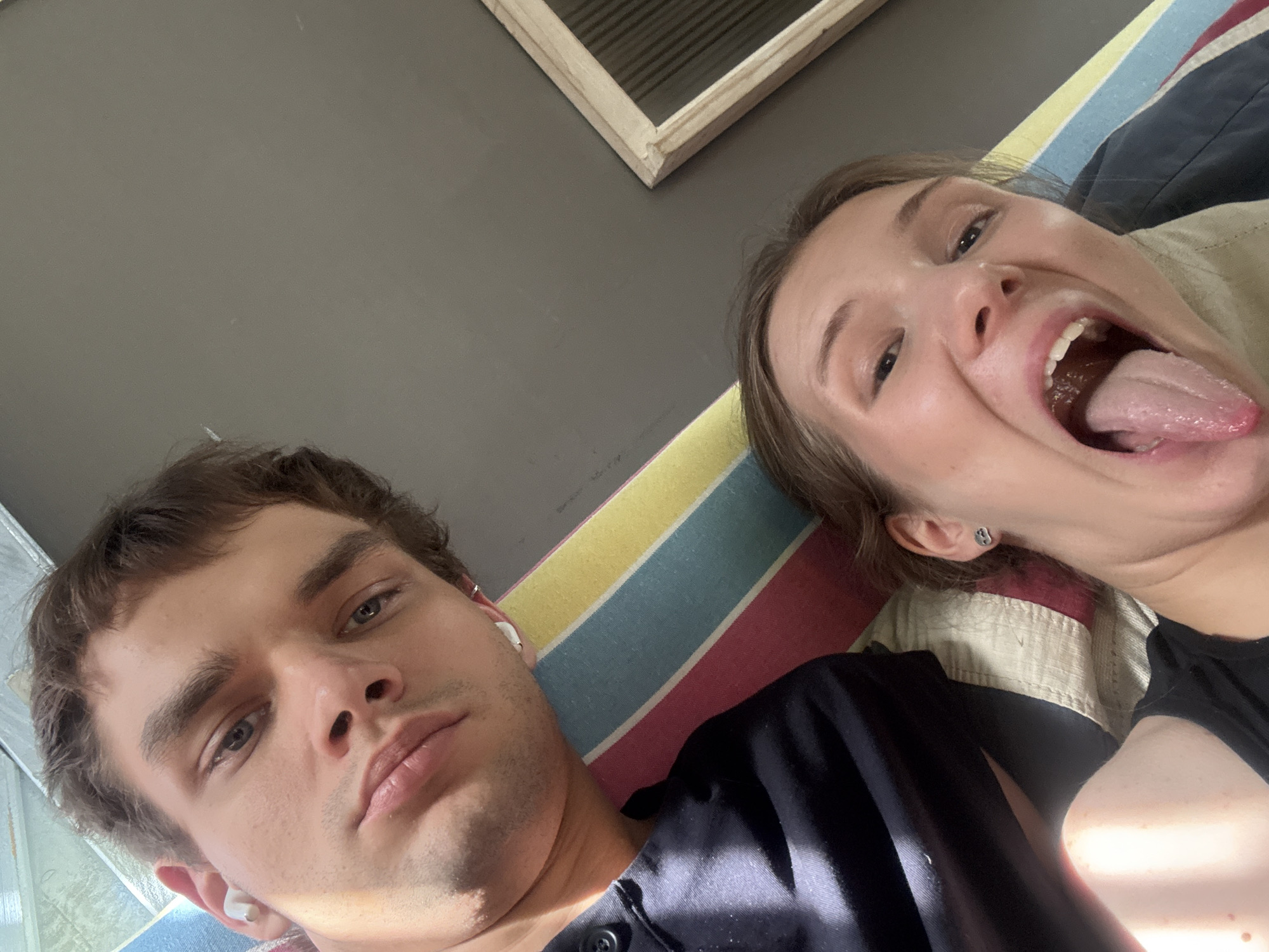Two individuals, a male and a female, are captured in an overhead shot while lying down, possibly taking a selfie. The scene is indoors, set against a plain grey ceiling or wall, with a wooden frame featuring horizontal slats visible in the upper right background.
The male is positioned on the left, looking directly towards the camera with a neutral, calm expression. He has short, dark brown hair and is wearing a dark-colored shirt. A white wireless earbud is visible in his right ear.
The female is on the right, displaying a playful and jovial expression with her mouth wide open and tongue sticking out. She has light brown hair and is wearing a dark top, with a lighter, possibly beige or cream-colored garment layered underneath or beside her. A small silver stud earring is visible in her left earlobe.
Both individuals are lying on a surface adorned with colorful stripes in shades of yellow, blue, red, and cream/off-white, which could be a blanket, pillow, or mattress cover. The lighting is bright and even, typical of an indoor setting during the day or under good artificial illumination. There is no visible text in the image. The photo depicts a casual, lighthearted moment. The location context provided is Putuo District, China.