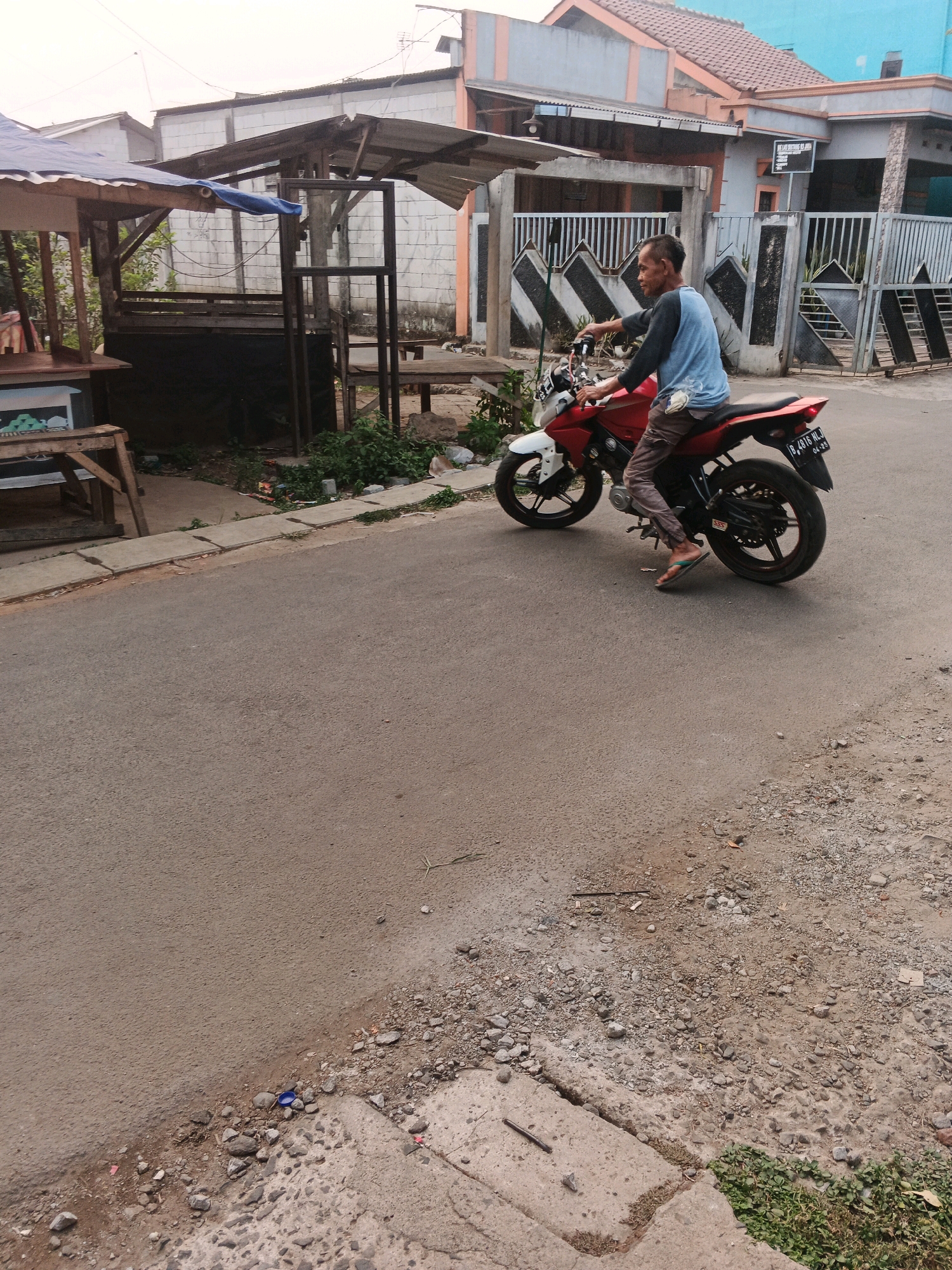 The image shows a man riding a red and white motorcycle in Kadu, Indonesia. The man is wearing a blue shirt and brown pants. He is riding on a paved road in front of a house with a blue and orange facade. There is a small wooden stall with a blue tarp to the left of the road. The weather appears to be overcast. The motorcycle has a license plate that reads "B 4816 HLJ". The man is looking to his right as he rides.