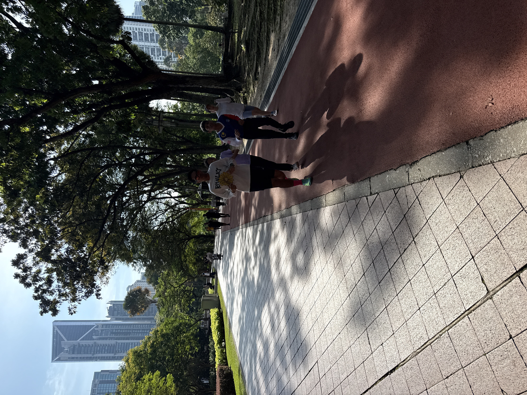 Three people are jogging on a red running track adjacent to a tiled pathway. The pathway is made of square gray tiles. The running track and the pathway are bordered by greenery, including trees and bushes. Shadows of trees are cast on the red running track. In the distance, high-rise buildings can be seen, indicating an urban setting. The building architecture indicates that it is in the city of Kuala Lumpur, Malaysia. A man is wearing a white long-sleeved shirt with a sunflower design on the front and black shorts with a pink sock. The person behind him is wearing a blue and white Malaysian jersey.