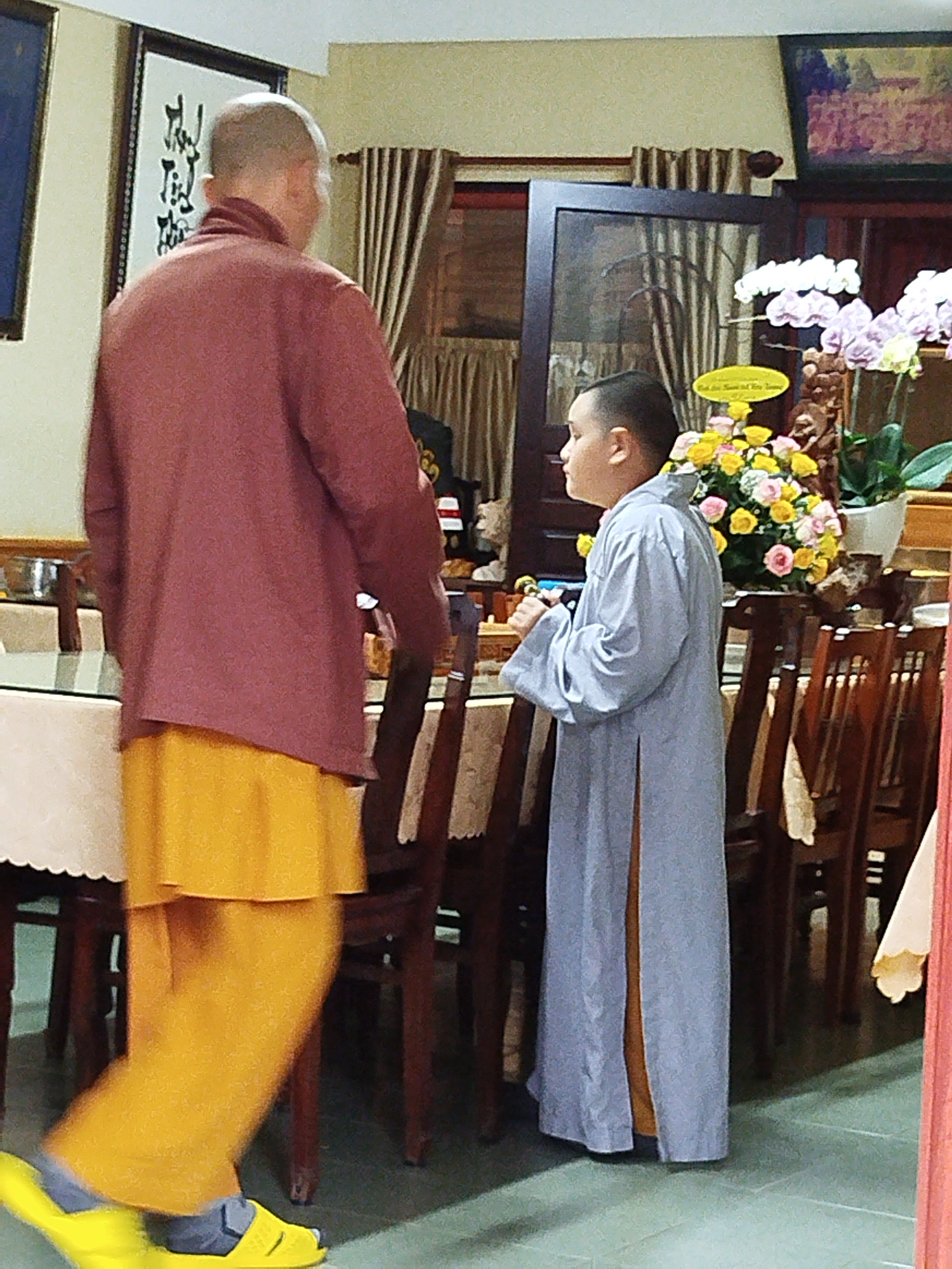 The image shows two individuals, likely Buddhist monks, in a room with furniture and decorations that suggest a religious or ceremonial setting. On the left, a person wearing a maroon robe and a bright yellow skirt is walking away from the camera. Their head is shaved. To the right, a younger person, also with a shaved head, is wearing a pale blue robe over a tan garment and is facing towards the left. They are holding what appears to be a small object in their hands. Behind them, a table is set with chairs, and a floral arrangement with pink and yellow flowers is prominent. A framed piece of calligraphy hangs on the wall. The flooring appears to be tiled. The overall impression is one of a quiet, possibly formal, gathering. There is no specific visual information in the image to indicate the location is Da Lat, Vietnam, or any specific city.