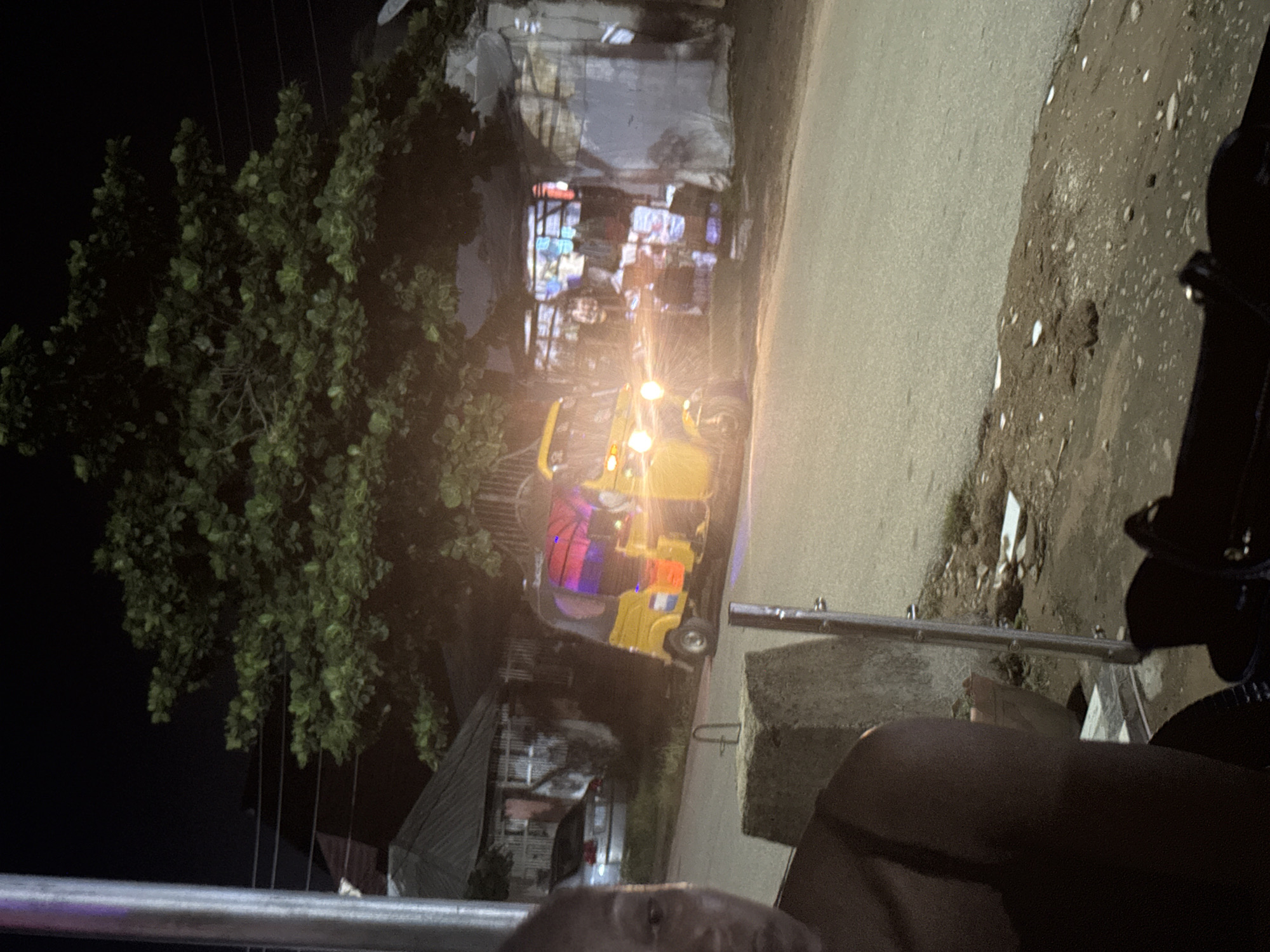 This image depicts a street scene at night in what appears to be an informal market area, possibly in Abuja, Nigeria. A yellow auto-rickshaw, commonly known as a "Keke Napep," is prominently featured in the center of the frame, its headlights illuminating the road and a nearby stall. The auto-rickshaw has colorful lighting inside its passenger area. To the left, a portion of a person's face and shoulder are visible in the foreground, suggesting the photographer is also present in the scene.
Behind the auto-rickshaw, several makeshift stalls are set up, covered by tarps and exhibiting various goods, some with colorful lights. The background reveals residential structures with corrugated metal roofs and simple facades, indicative of a less affluent neighborhood. A silver car is parked to the left of the auto-rickshaw, partially obscured by a tree. The environment appears to be a paved road with a dirt shoulder, littered with debris. A large tree with dense foliage overhangs the scene, casting shadows and contributing to the nighttime atmosphere. The overall impression is that of a bustling, if somewhat unorganized, commercial hub operating after dark.