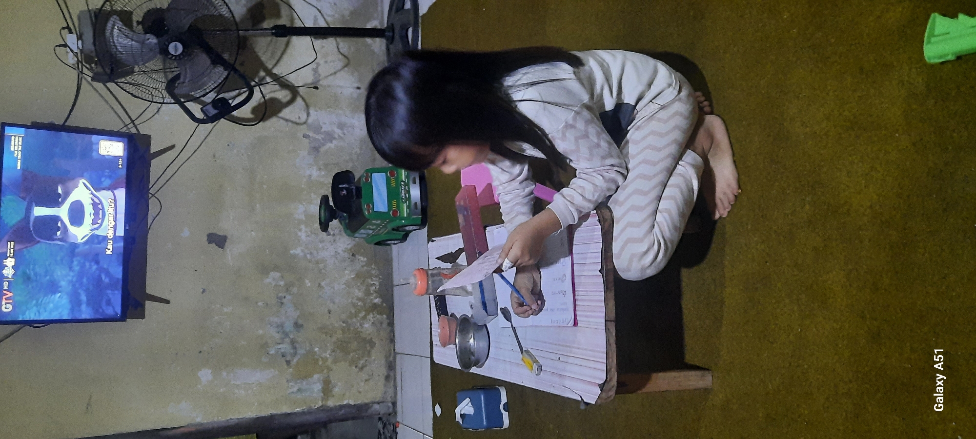 In Ploso, Indonesia, a young girl with long black hair, wearing a white and beige zig-zag patterned loungewear set, is seated on a yellow-brown carpet. She is squatting beside a low, light-colored wooden table, actively engaged in writing on a piece of paper with a blue pencil. In front of her on the table are various items, including a small metal bowl, a tissue box, a bottle with an orange lid, and what appears to be a pink pencil case or box. To the left of the table, a blue tissue dispenser is visible. In the background, a green toy truck with "GARBAGE" written on it is placed on the floor. A large, oscillating fan stands to the right of the scene, and an old, textured wall with some discoloration serves as the backdrop. Above the wall, a television screen displays a cartoon image of a dog with the text "Kau dengar itu?" and "B-15+" visible on the screen. The lighting in the room appears to be dim, contributing to the overall atmosphere.