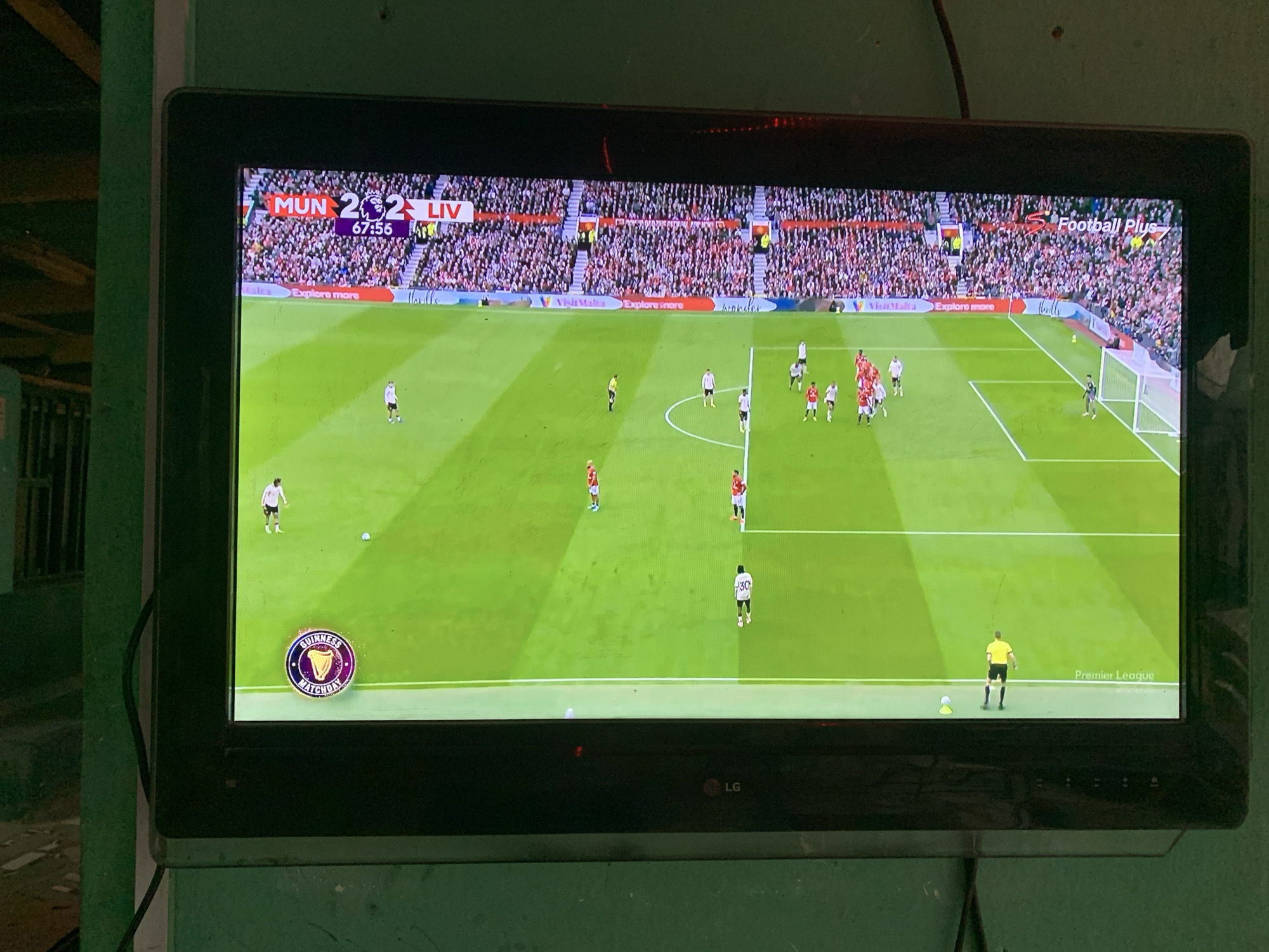 The image depicts a television screen displaying a live broadcast of a football match between Manchester United (MUN) and Liverpool (LIV), with the score tied at 2-2 and the time at 67:56. The match is part of the Premier League. A "Guinness Matchday" logo is visible in the bottom left corner of the screen. On the pitch, players are positioned in a defensive formation near the penalty box. The broadcast is airing on "Football Plus". The TV is an LG. The location may be New Stadium, Nigeria, but it is not visible in the image.