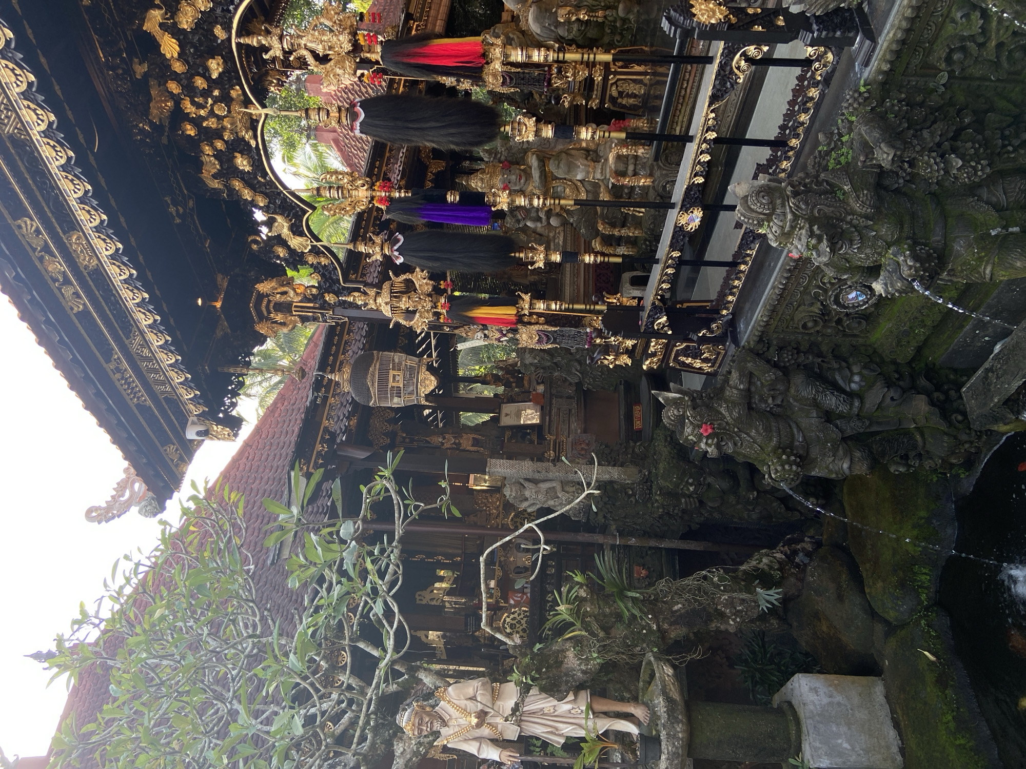 The image depicts a courtyard in what appears to be a Balinese temple complex. The location is most likely within the Ubud District, Indonesia, given the architectural style and cultural artifacts present. The architecture is characterized by intricate carvings, detailed woodwork, and ornate decorations, many of which are gilded. Statues and shrines, some adorned with moss and vegetation, are placed throughout the space. A tiered structure with a dark roof, likely a pavilion or shrine, features elaborate gold-colored embellishments. Prayer poles are visible with colorful adornments hanging. A small pond or water feature is visible, with stone carvings serving as spouts for water. Plants and trees add a natural element, contributing to a sense of a tranquil sanctuary. The scene suggests a place of religious or cultural significance and traditional practices. The image is taken from a low vantage point, looking upwards and across the courtyard.