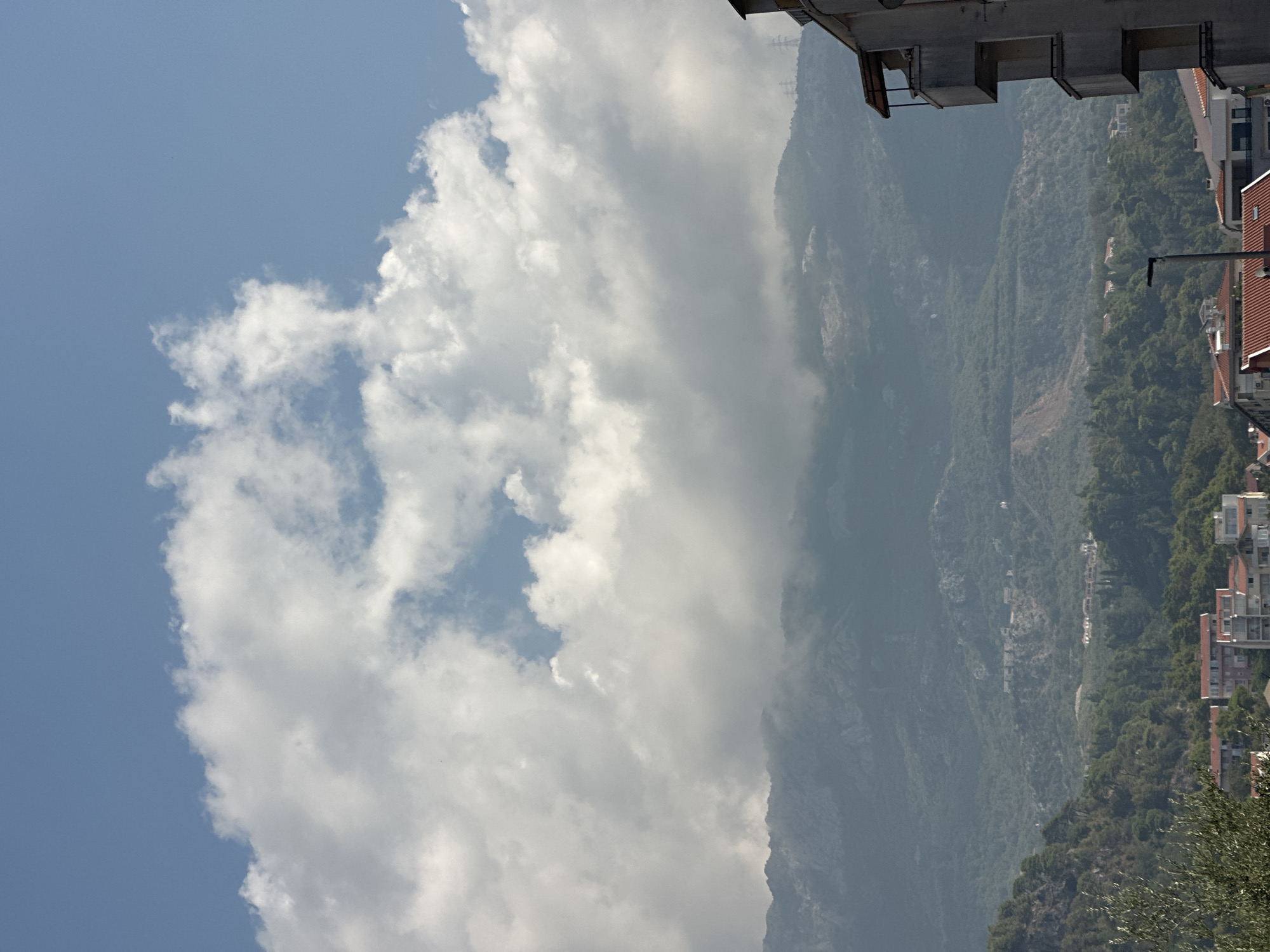 A view of mountains partially obscured by clouds is visible in the background of this image, taken in Budva, Montenegro. In the foreground, a portion of a building with balconies is on the right side of the frame. Below the mountains, a town with red-tiled roofs and buildings can be seen nestled amongst green trees. The sky is blue with scattered white clouds.