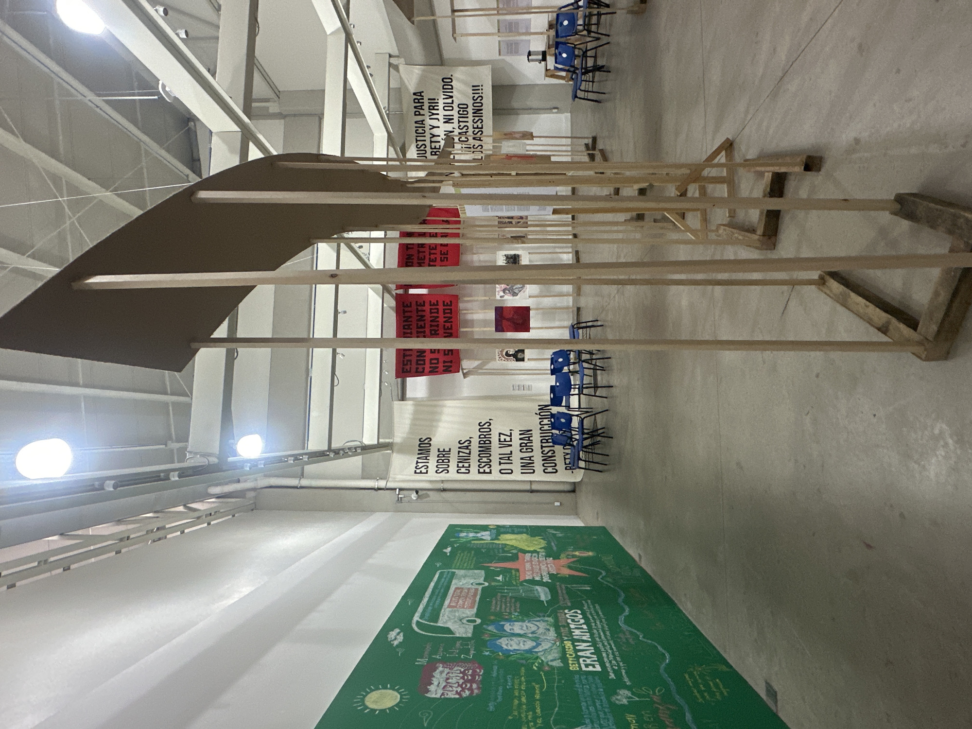 The image shows an indoor exhibition space in San Bernardino Tlaxcalancingo, Mexico. The foreground features a wooden structure, partially obscuring the view of several banners and posters displayed further back. One banner reads "JUSTICIA PARA BETTY Y JYRI! NI PERDON, NI OLVIDO. CASTIGO A LOS ASESINOS!!!" Another reads "ESTAMOS SOBRE CENIZAS, ESCOMBROS, O TAL VEZ, UNA GRAN CONSTRUCCIÓN". A green wall exhibit is visible on the left side of the frame, showing the text "ERAN AMIGOS" near the bottom. Blue plastic chairs are scattered in the background. The space has high ceilings with exposed support beams.
