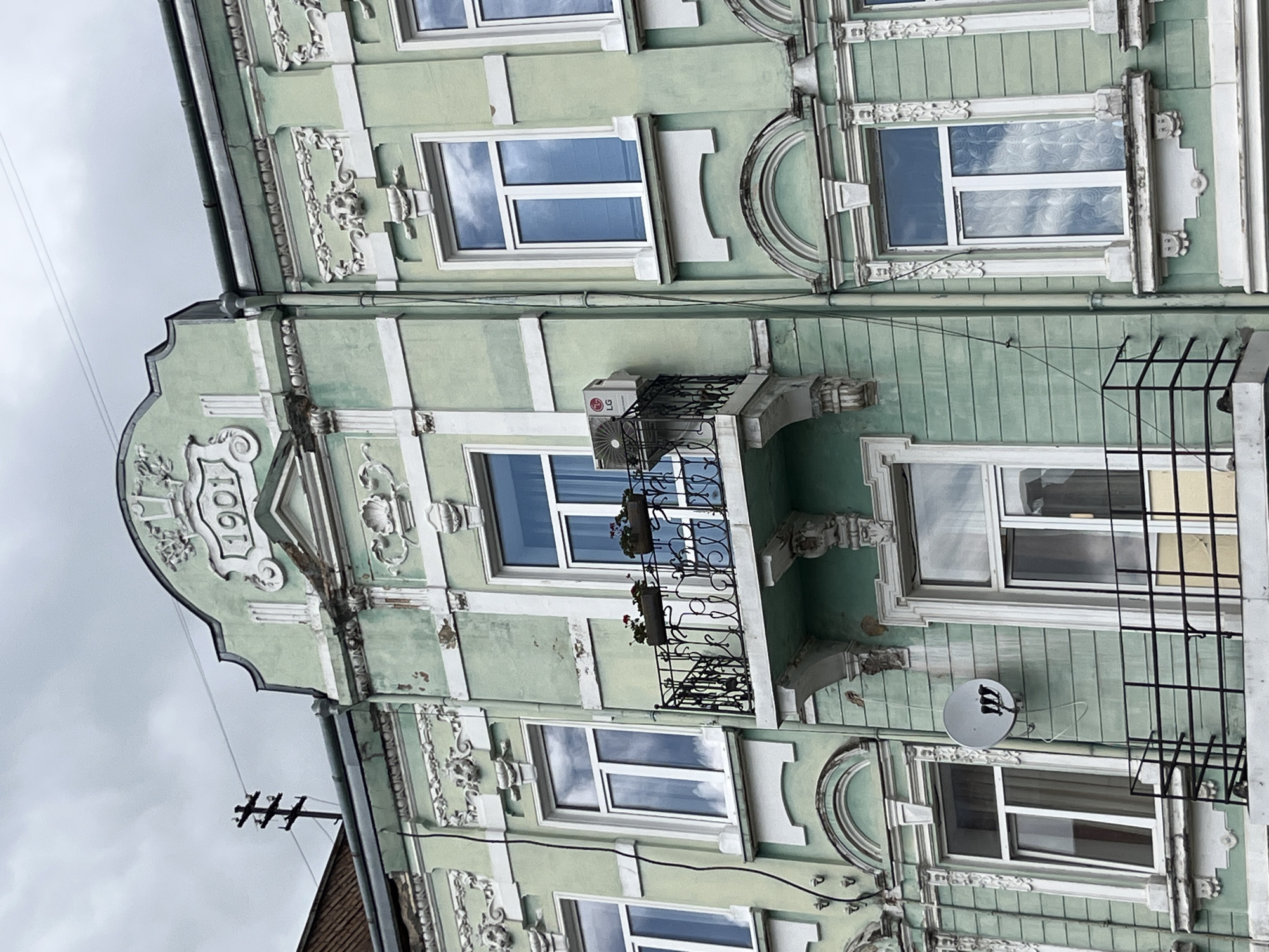 This is a medium shot, eye-level, exterior, daytime photograph of a pale green building with ornate white architectural details and a pediment dated "1901" at the top. The building is located in Drohobych, Ukraine. The facade features multiple windows, some with decorative balconies. An air conditioning unit is mounted to one balcony. A satellite dish is mounted to the wall to the left of a window. The sky is overcast.