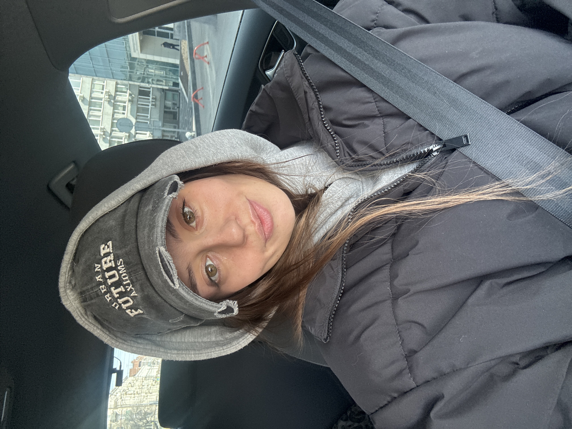 A young woman is captured in a casual, selfie-style photograph taken inside a car, likely from the passenger seat, with an urban background visible through the window. She looks directly at the camera with a subtle, pleasant smile. Her light brown hair frames her face, and her eyes are a distinct brown or hazel.
She is dressed in a dark grey or black puffer jacket worn over a grey hooded sweatshirt, with the hood up. A distressed grey baseball cap rests on her head, adorned with white embroidered text that reads "URBAN" above "FUTURE" and "AXIONS" (or a similar word) below it. A dark seatbelt crosses diagonally over her chest and left shoulder.
Through the car window, tall, modern buildings with numerous windows and visible air conditioning units indicate an urban environment. Red, curved metal barriers can also be glimpsed outside, partially obscured. The lighting suggests it is daytime, with soft, even illumination, possibly indicating an overcast sky. The overall scene conveys a candid moment in a city setting. Given the provided context, this image depicts a scene in Krasnoyarsk, Russia.