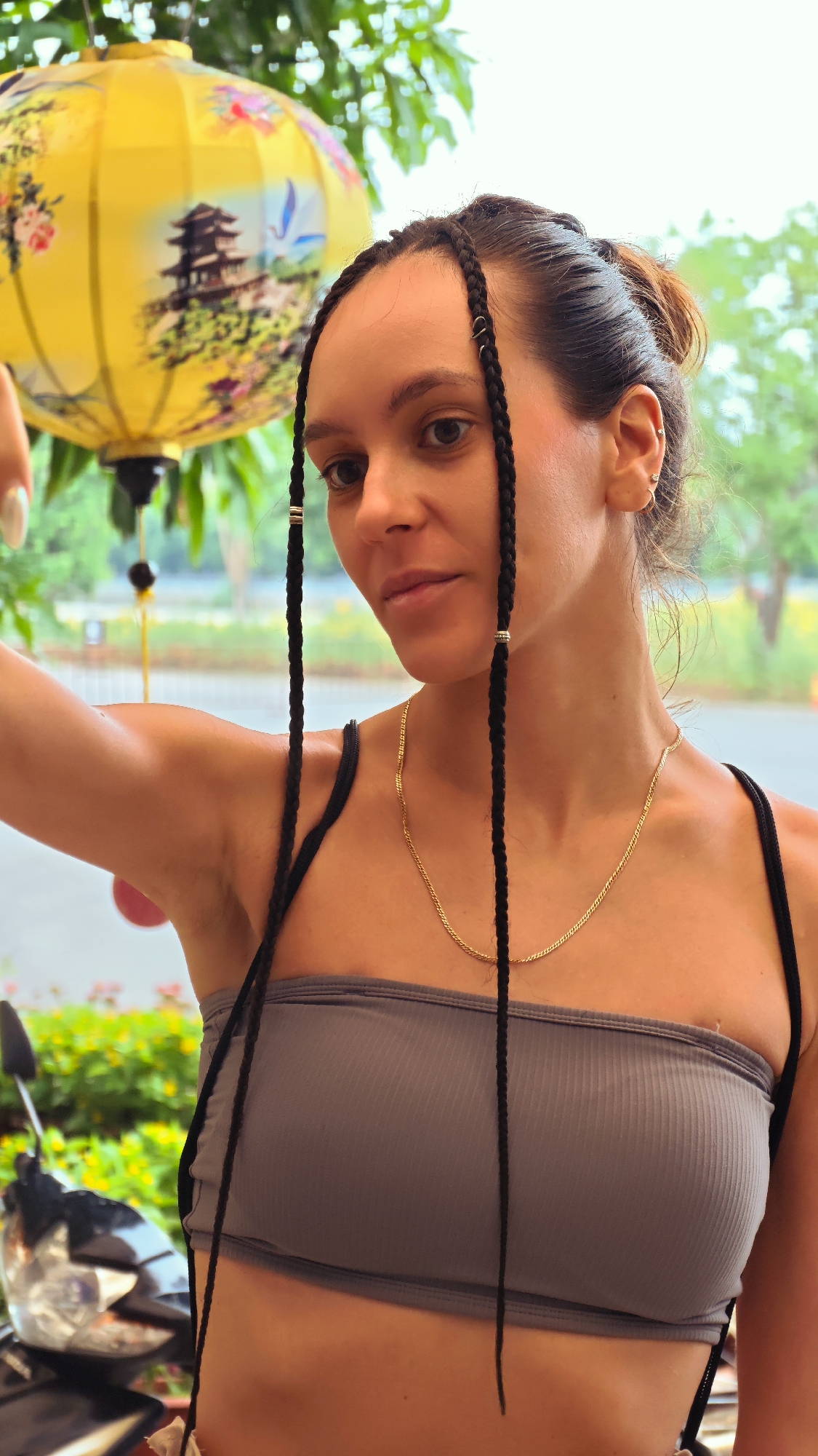 A medium shot captures a young woman with dark braided hair, her head turned to the right and looking down. She wears a grey ribbed tube top and a gold chain necklace. Her braids, adorned with small metal rings, hang down in front of her.
Behind her, a large yellow lantern, decorated with intricate paintings of a pagoda and flowers, hangs from above. The background is softly blurred, showing greenery and hints of what appears to be a street scene with a motorcycle parked to the left. The lighting suggests it is daytime, possibly overcast, with diffused natural light. The overall impression is a candid moment, perhaps taken outdoors in a vibrant, possibly Southeast Asian, setting like Tây Hoa Lư Ward, Vietnam, given the context provided. The woman's expression is neutral, her gaze directed downwards, giving a thoughtful or introspective feel to the image.